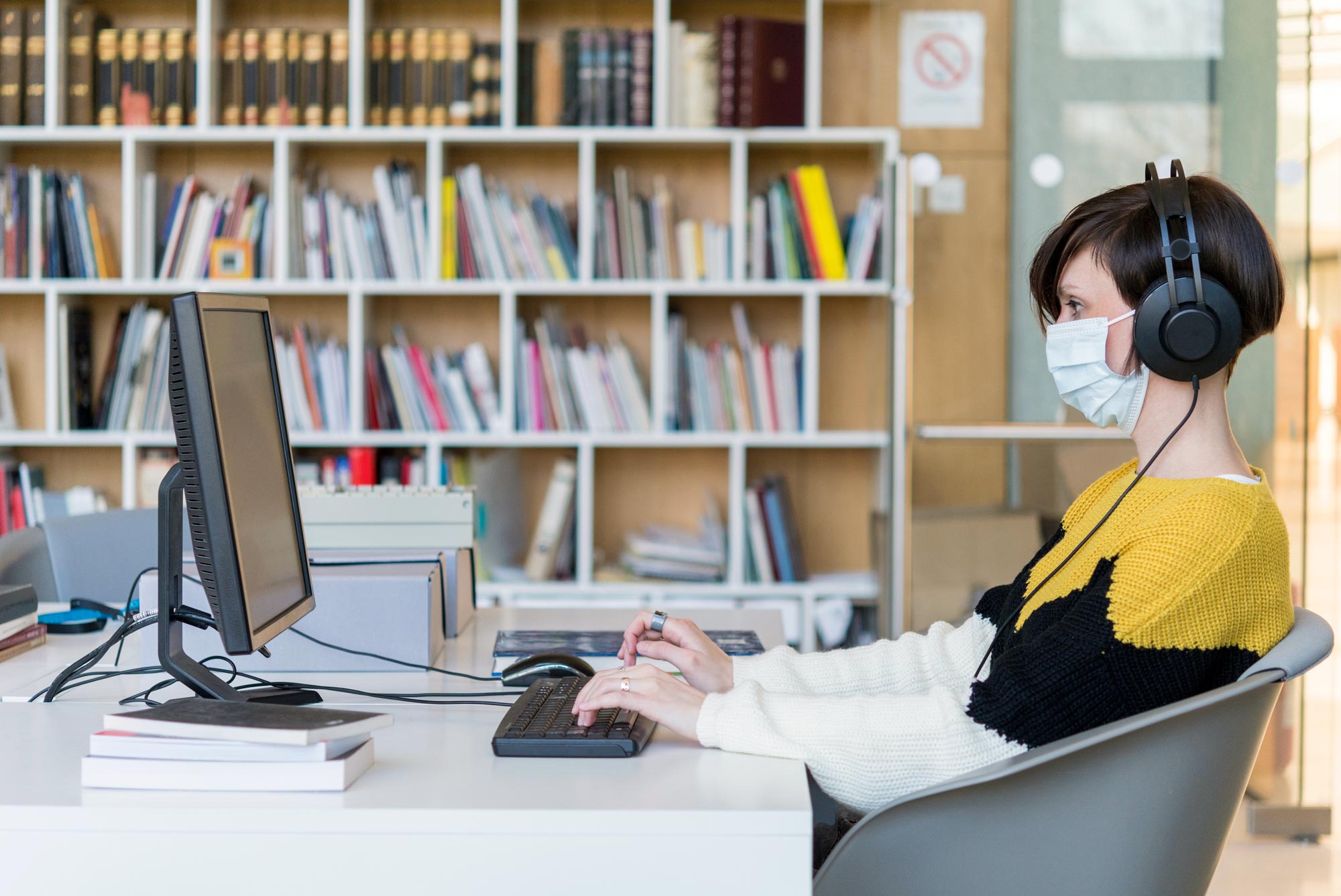 Woman sitting at a desktop computer wearing headphones and a mask