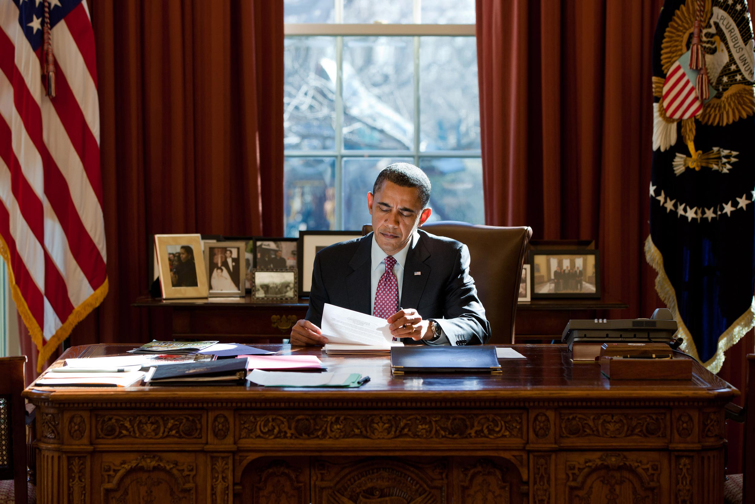Barrak Obama sitting at the Oval Office desk reading a piece of paper