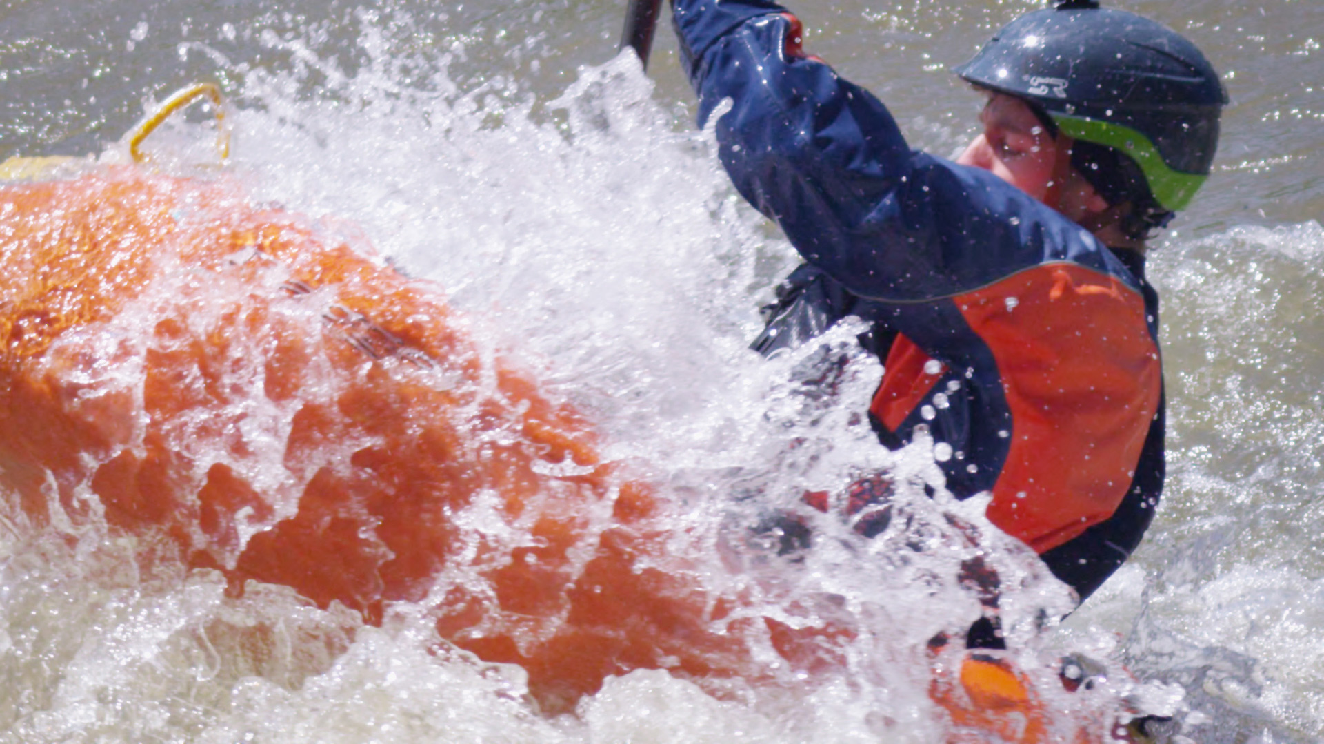 Man white water rafting.  Water is splashing up over him and as he is paddling