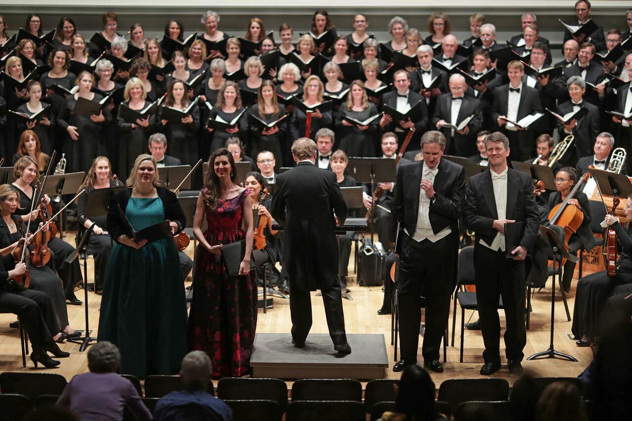 From left to right, Christina Pier, Adelaide Muir Trombetta, Michael Slon, and Mark Mowry and David Newman stand in front of an orchestra and the rest of the choir