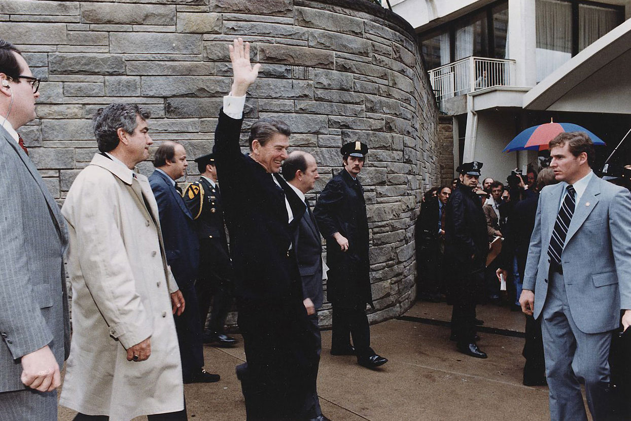 President Reagan raising his hand in the air as he walks passed a crowd with his security detail