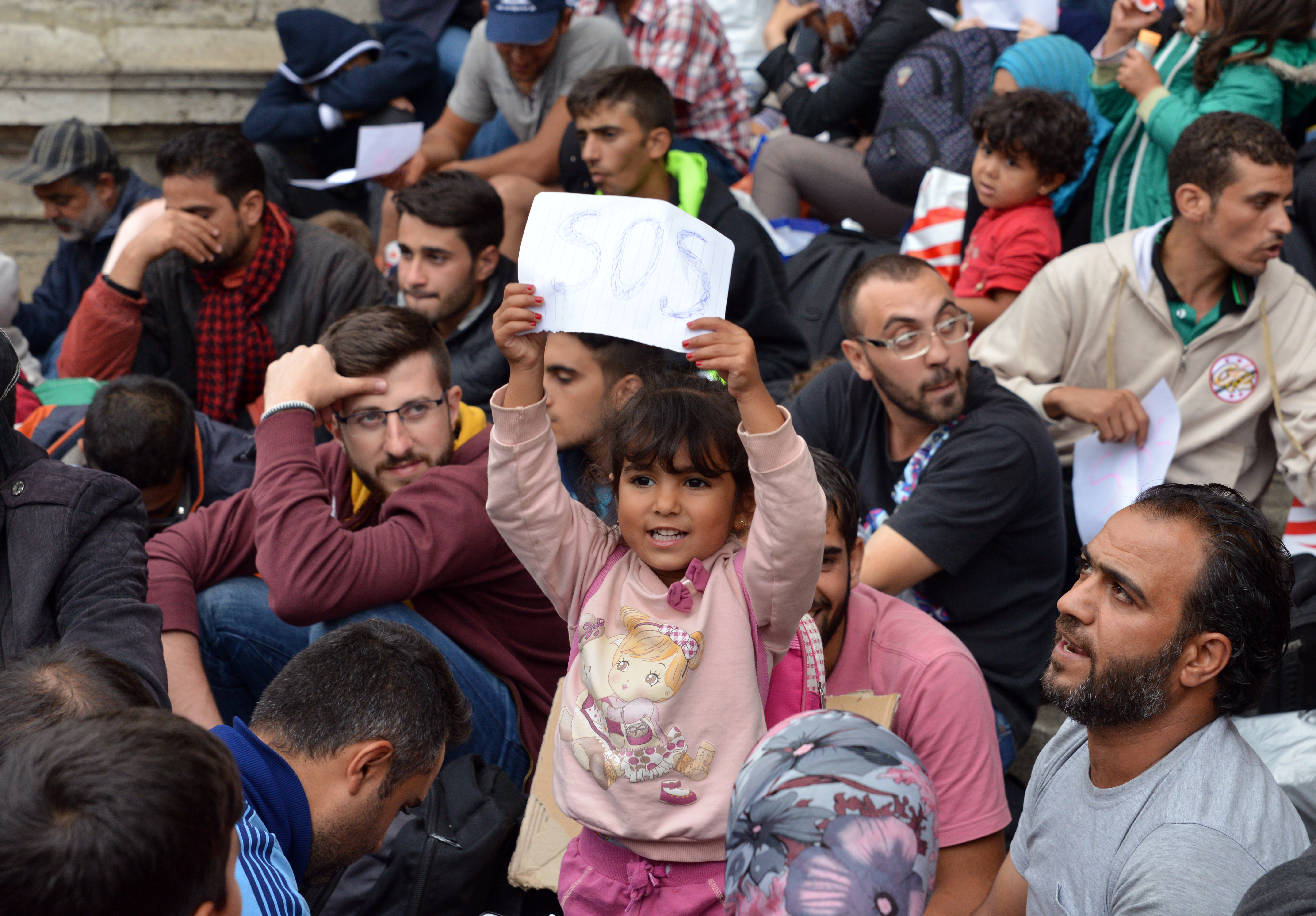 Group of people gather on the ground while a little girl holds up a piece of paper that reads sos