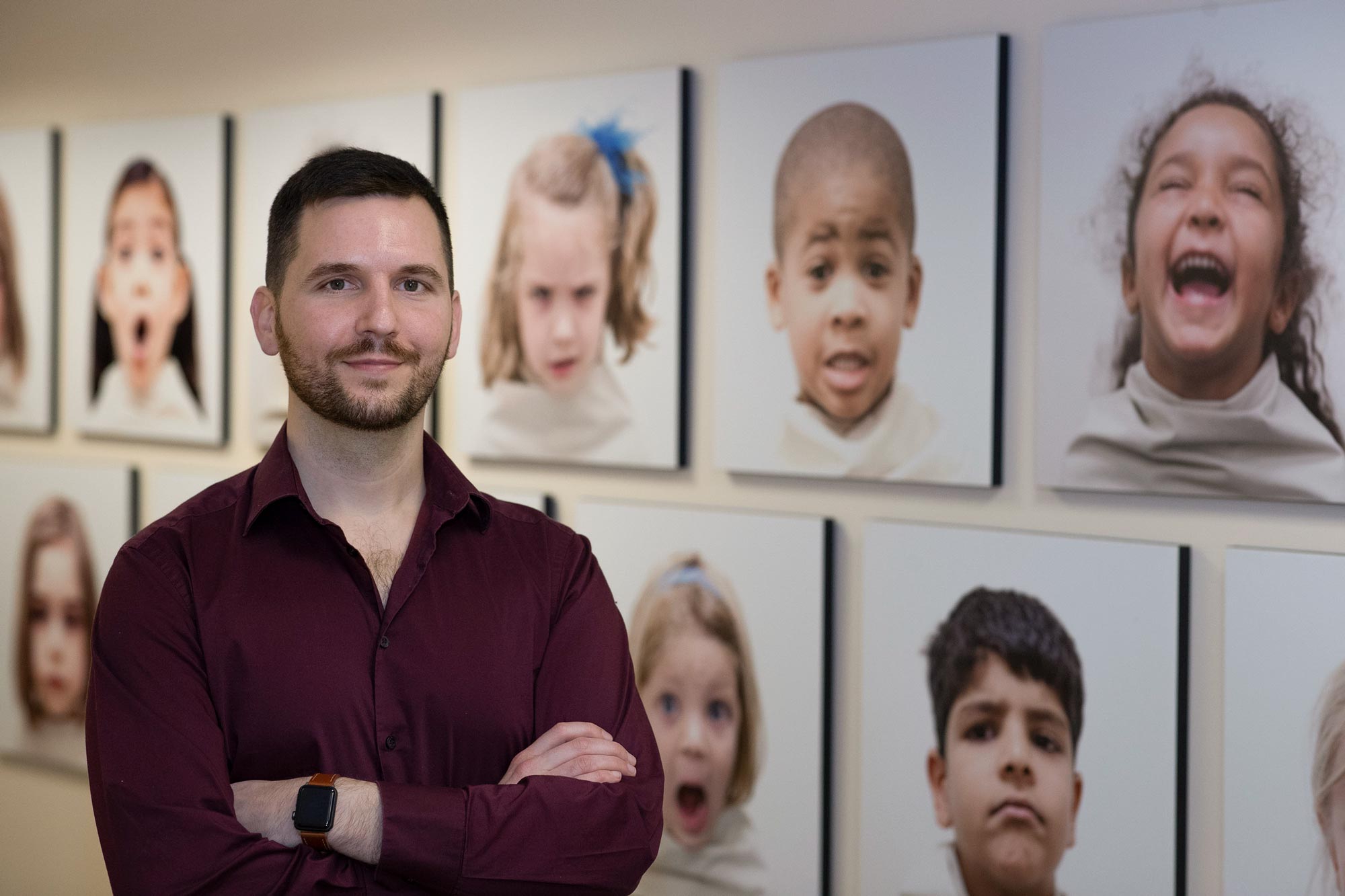 Stefen Beeler-Duden stands in front of portraits of children with his arms crossed looking at the camera