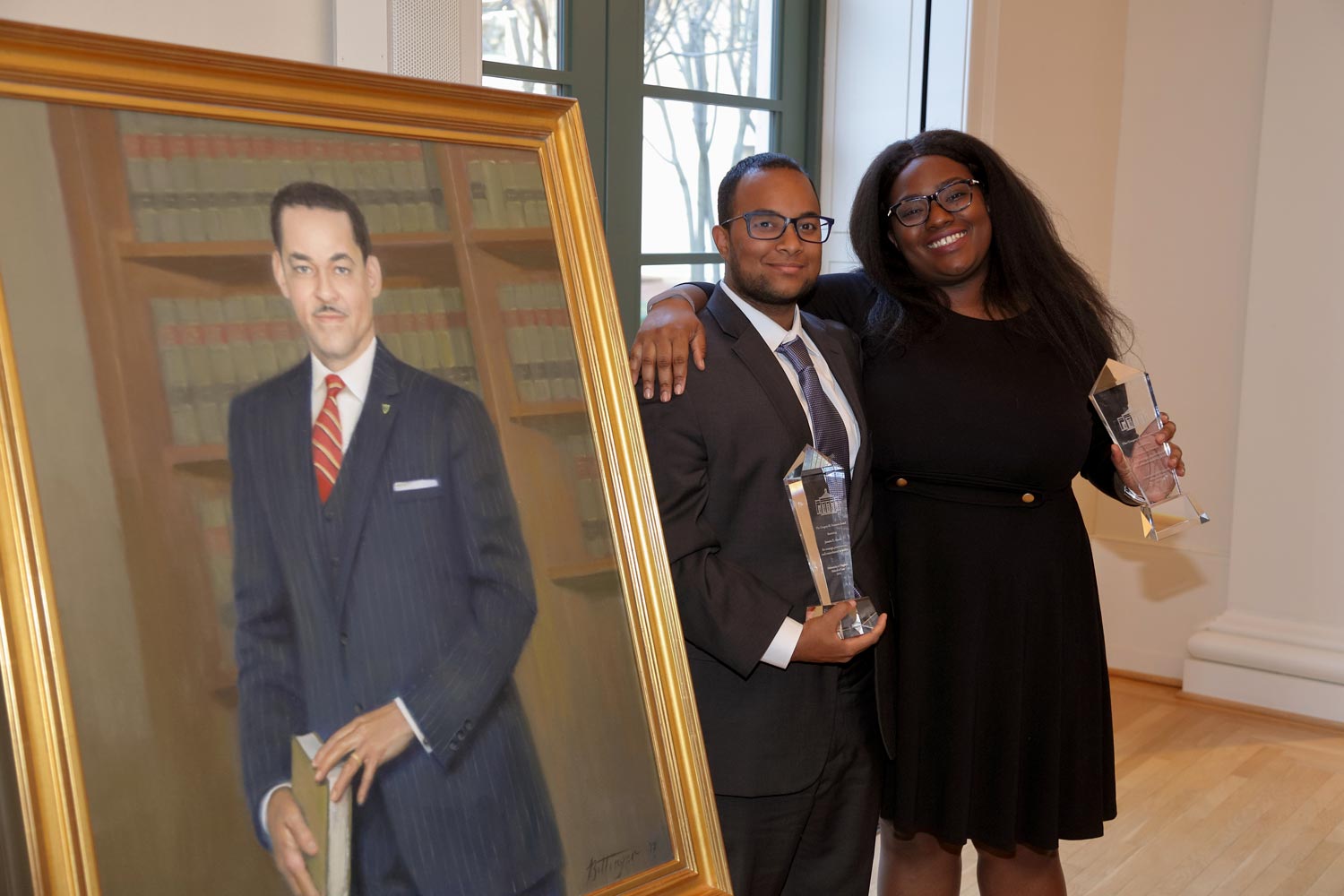 Jamaica Akande and Toccara Nelson stand next to a painting of Gregory H. Swanson holding their glass awards