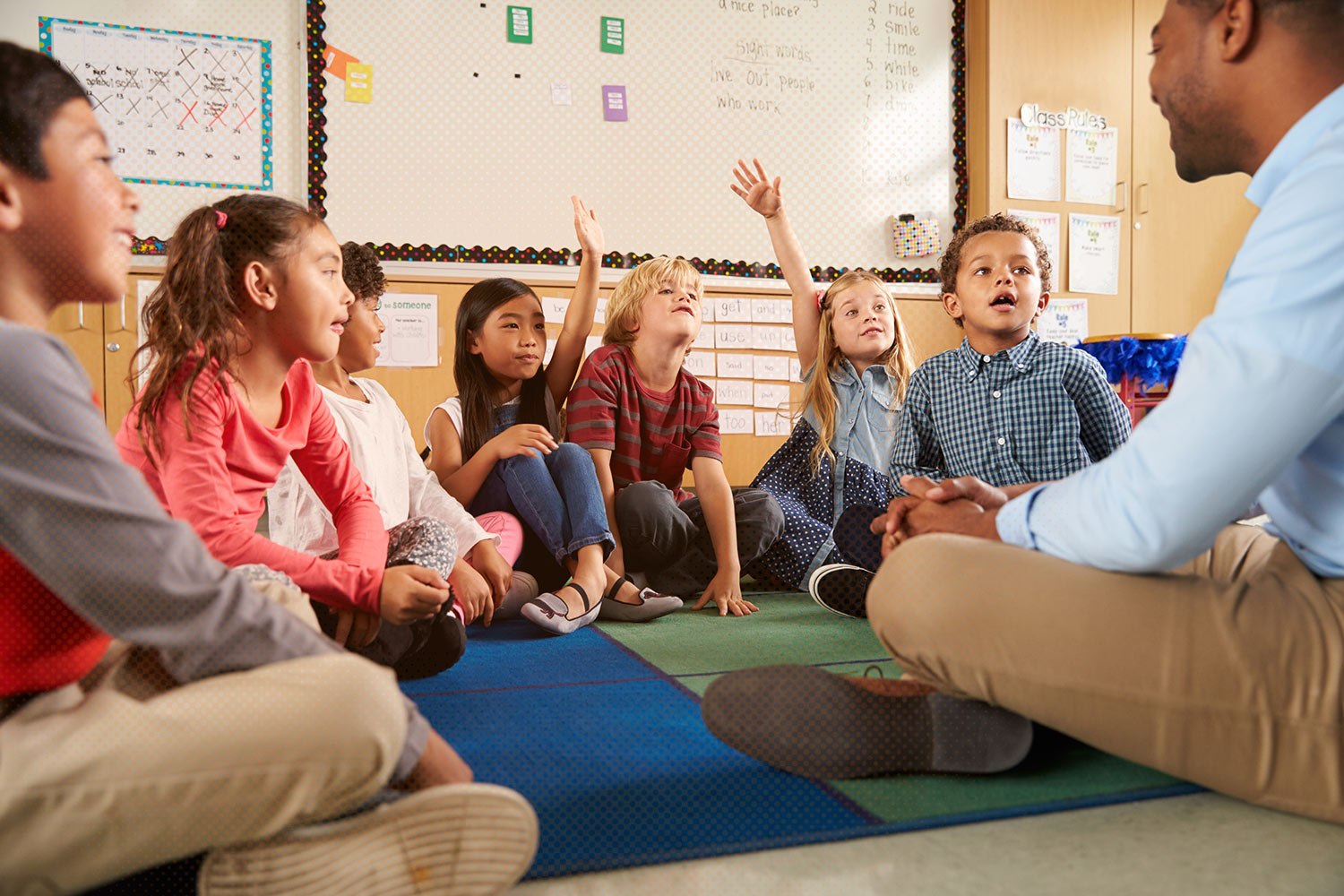 Teacher sitting on the floor with children raising their hands