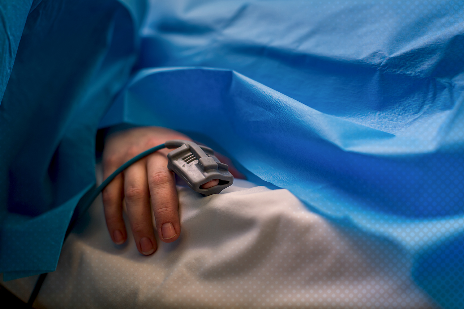 Patients hand on a bed sticking out of a blue surgical sheet with an oximeter 