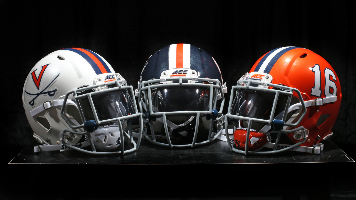 Three UVA football helmets sit on a table.  Left to right: White helmet, UVA Blue helmet, UVA orange helmet