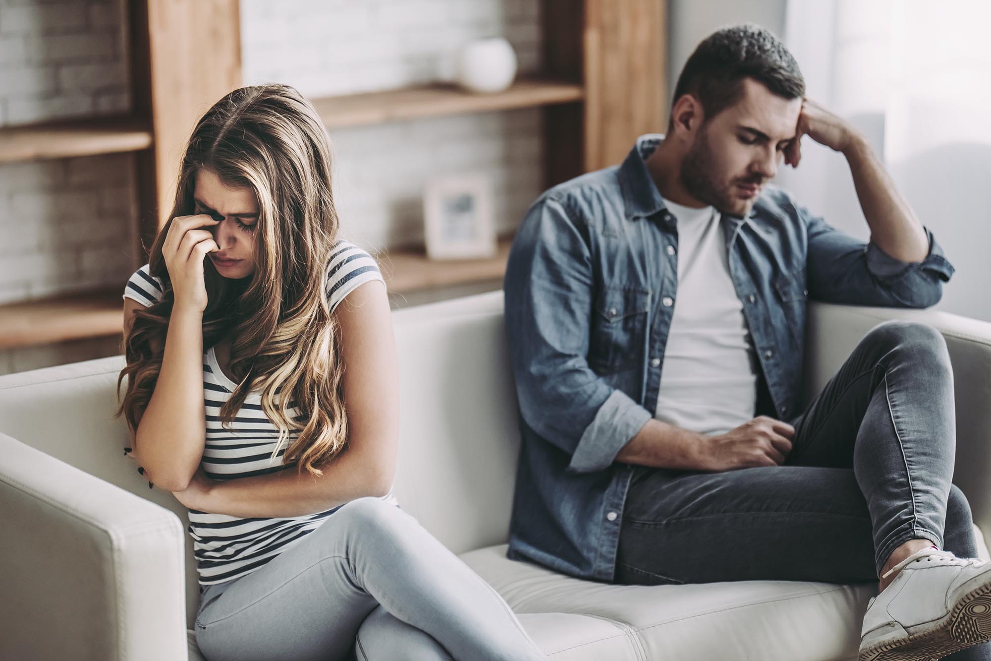 Two people sitting on opposite ends of the couch both upset.  Woman, left, rubbing her eye