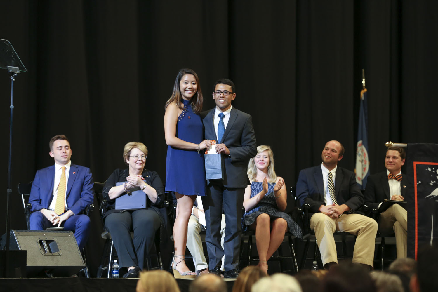 Ngan K. Pham, left, Mario J. Sukkar, right, stand together smiling at the camera on stage as Ngan hands Mario a blue gift bag