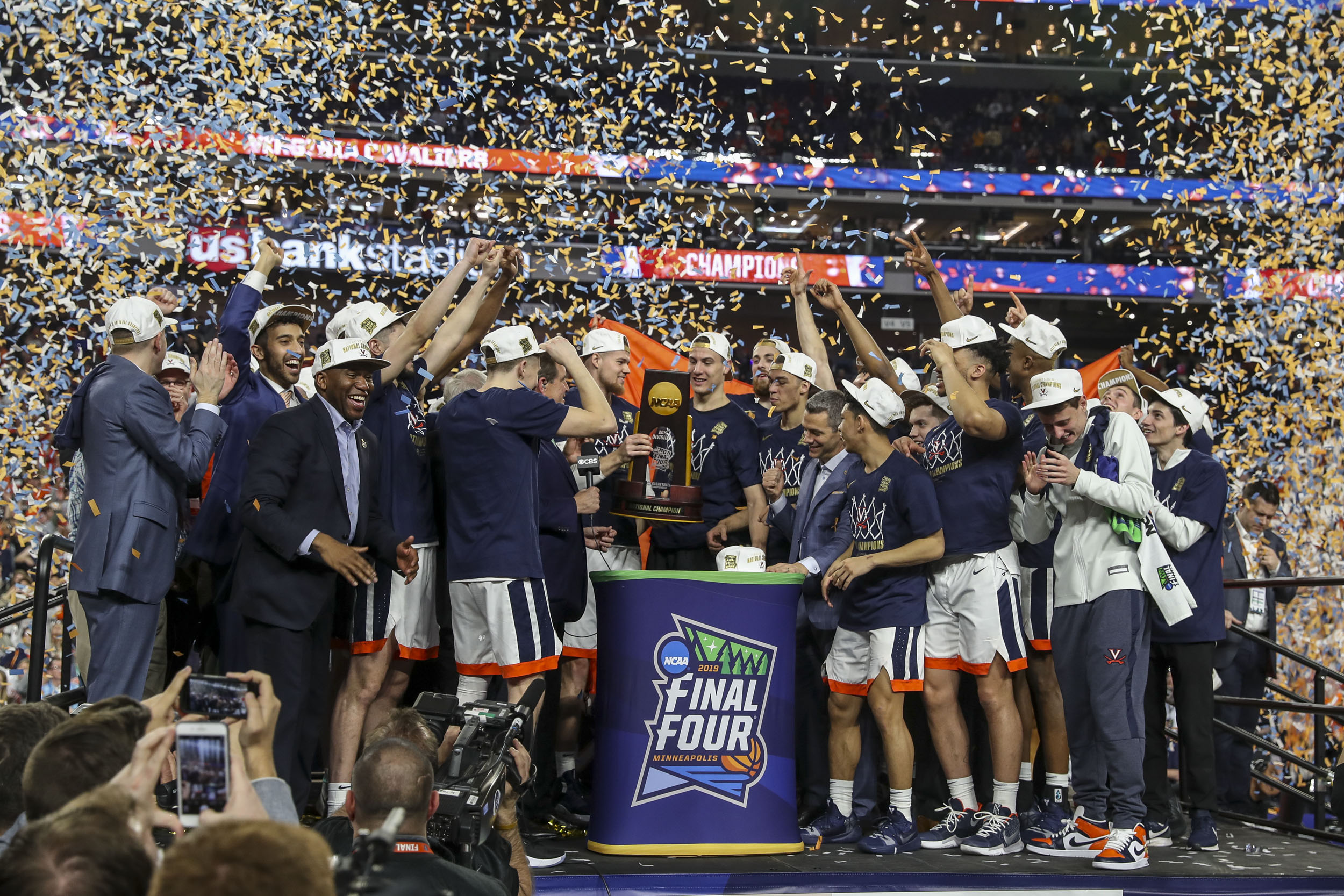 Mens Basketball team on stage holding the NCAA trophy while confetti falls from the sky