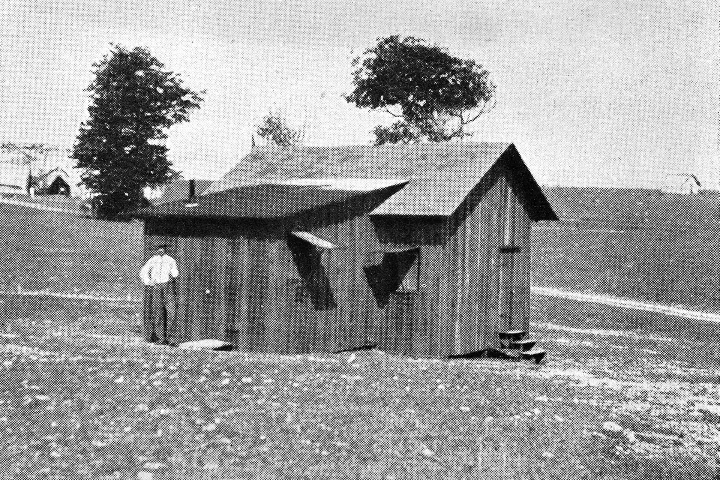 Black and white photo of a man standing outside of a small wooden house