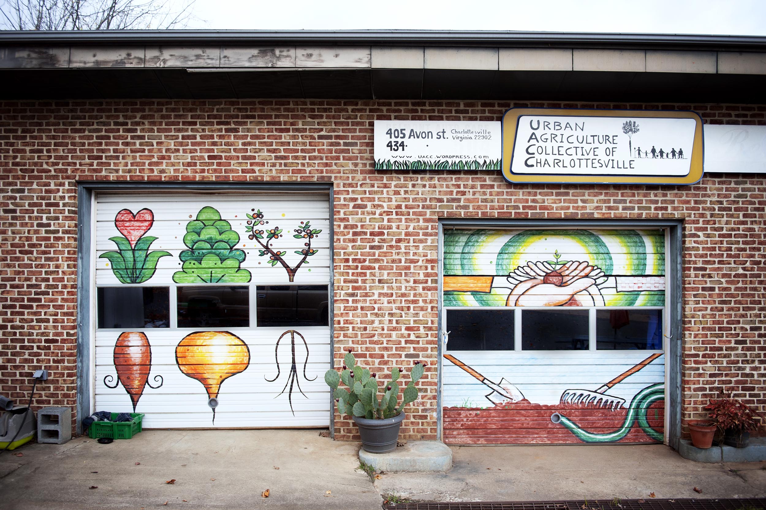 Garage doors on a brick building with paintings.  Left: 3 vegetables right: hands holding a vegetable and a shovel, rake, and water hose sitting on dirt