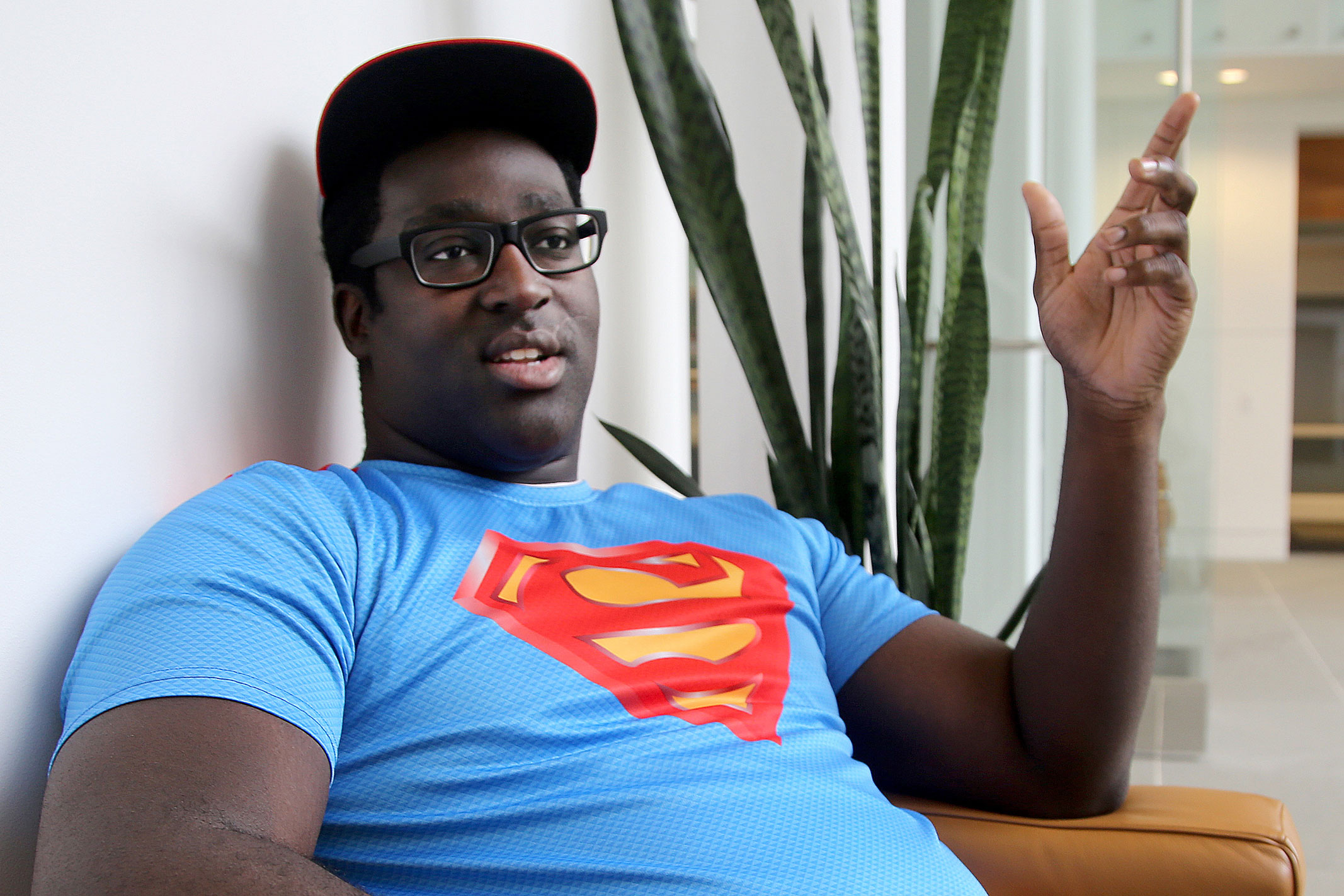Man sitting in a tan chair talking wearing a superman shirt