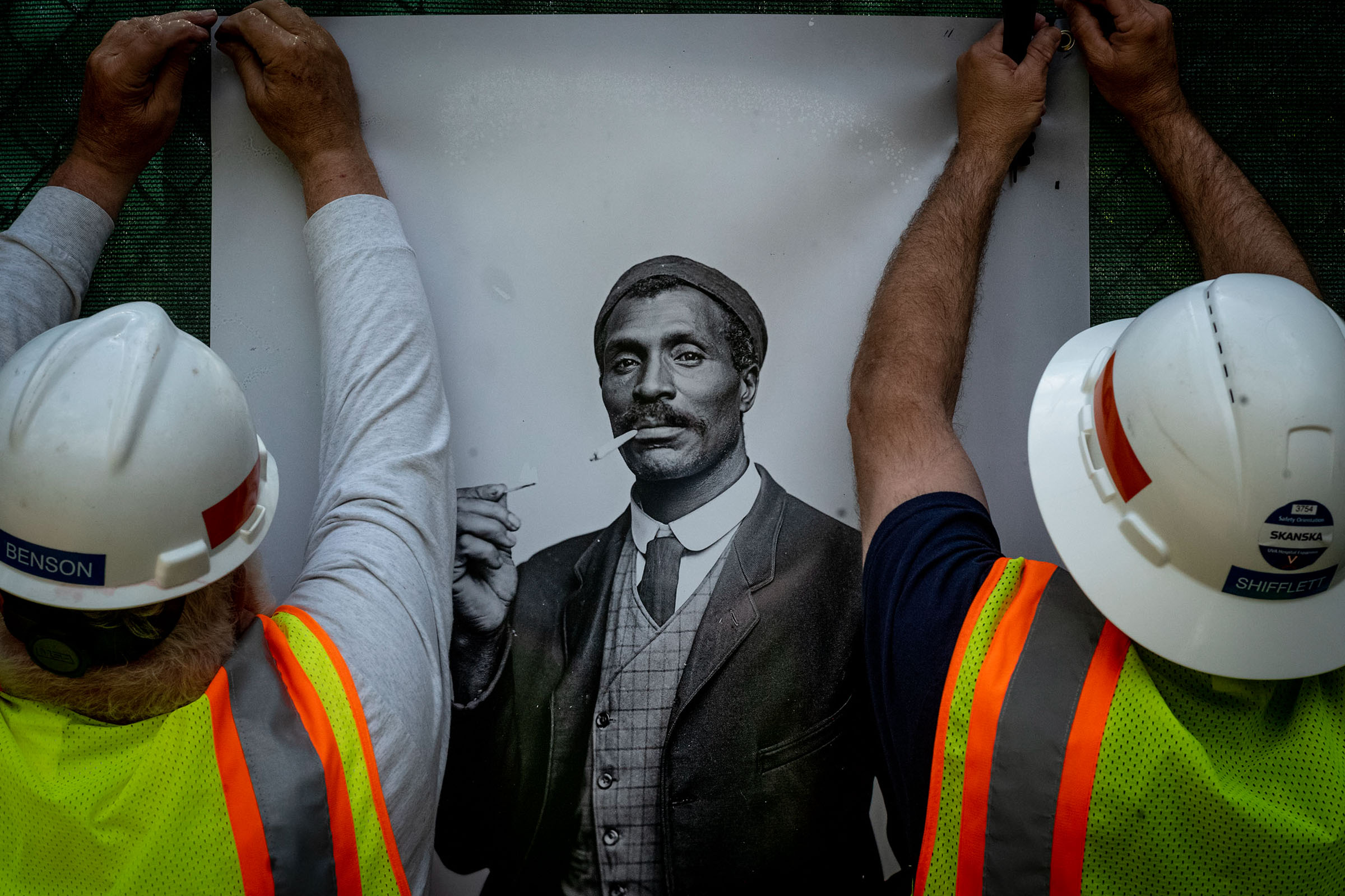 Construction worker handing up a poster of a black man dressed in a suite smoking a cigarette 