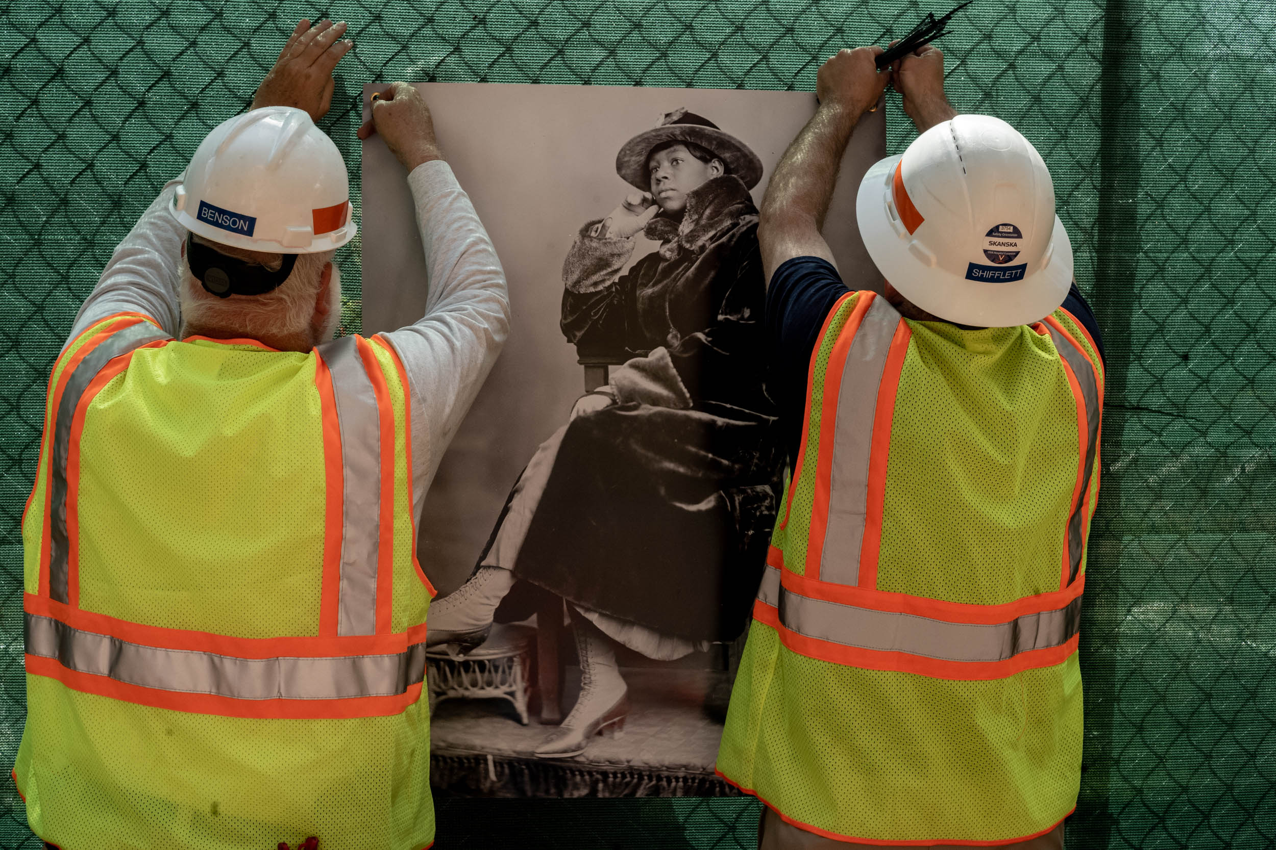 Construction workers hang images on a chain link fence