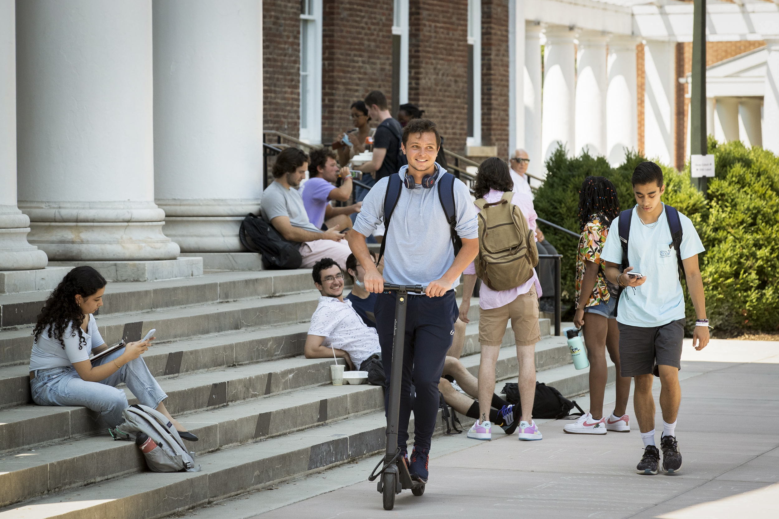 Students sittiing on steps, walking, and riding a scooter on a sidewalk in front of the steps