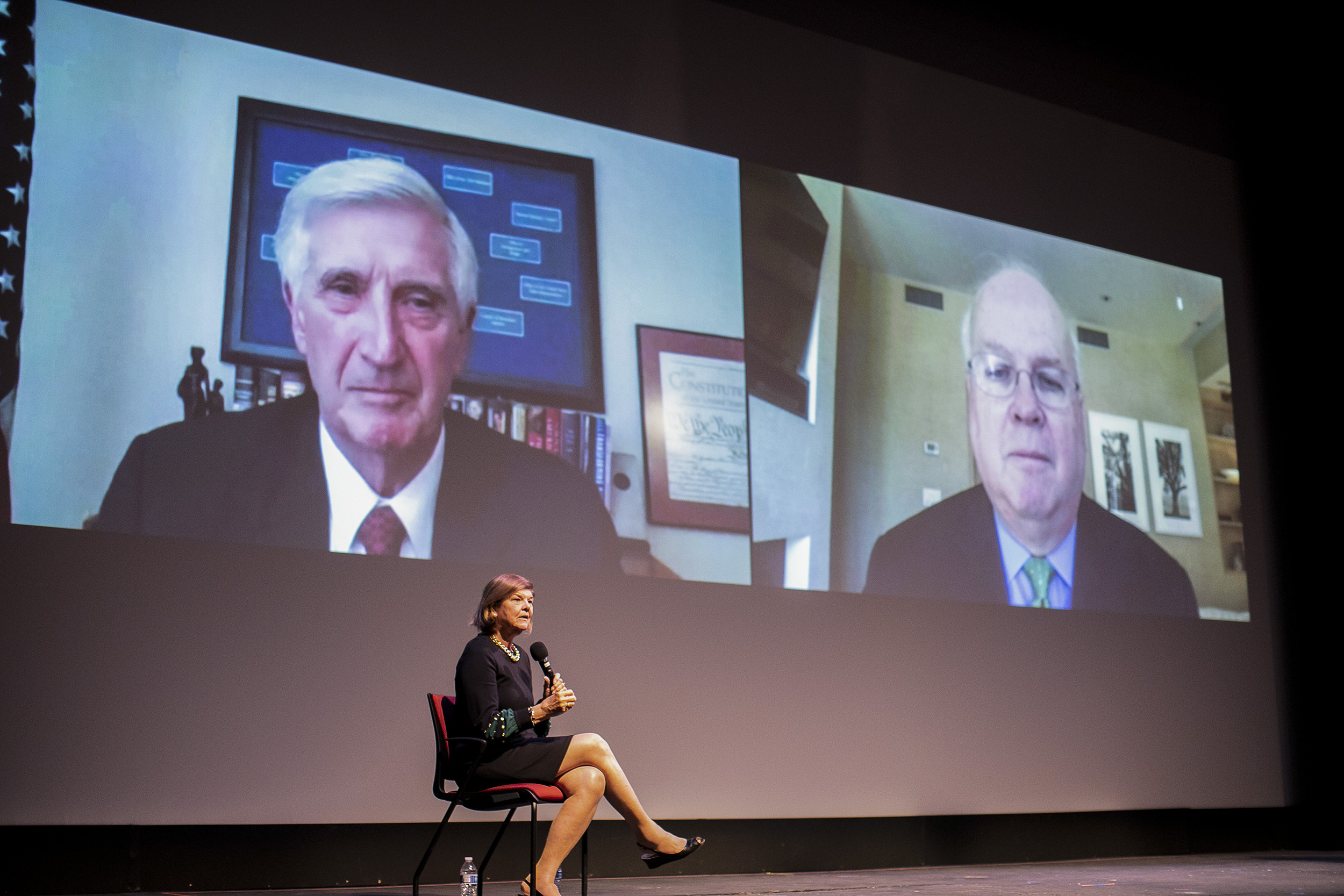 Woman on stage speaking while two men on a split zoom screen listen