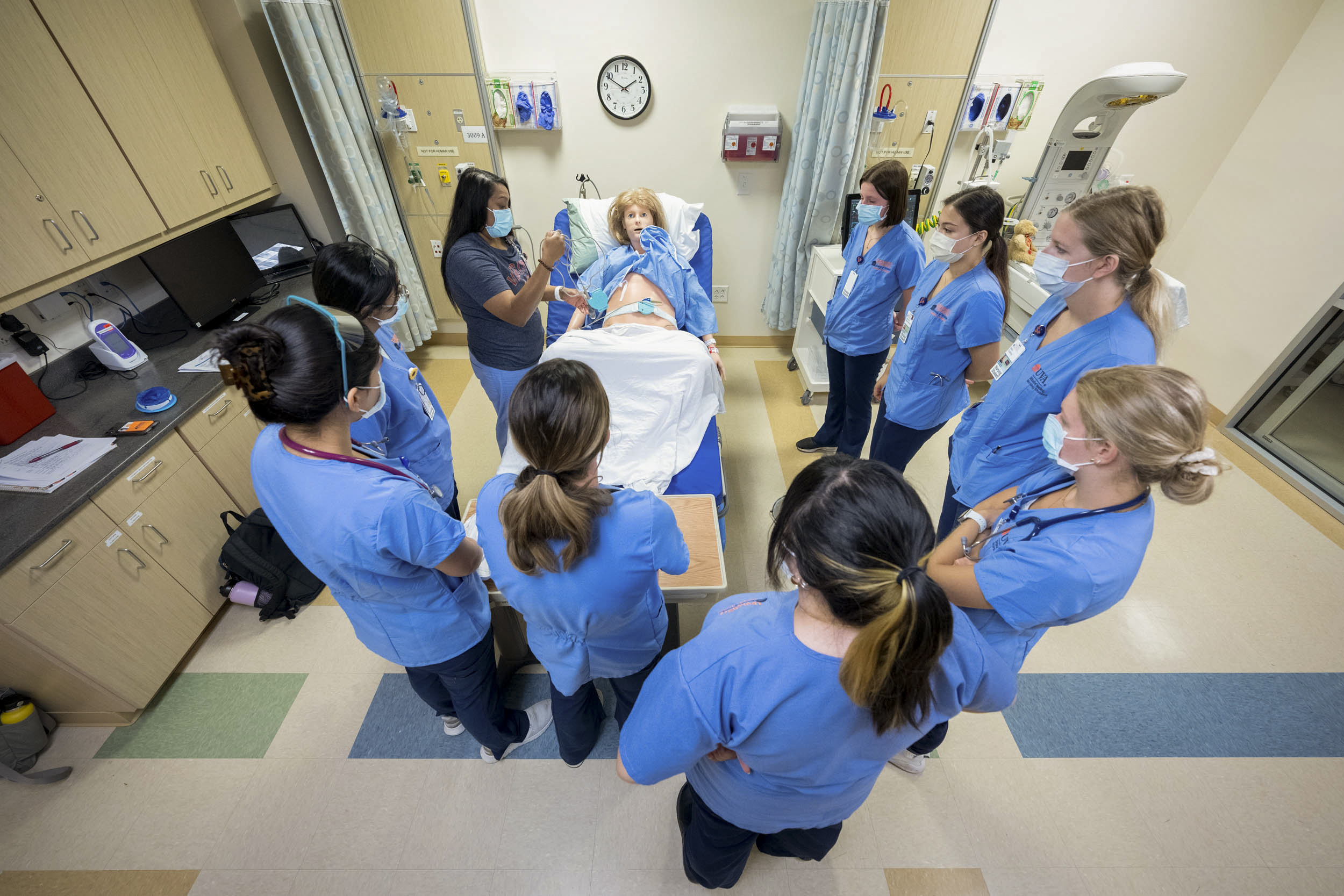 Nursing students stand around a fake human body in a bed listening to a RN speak