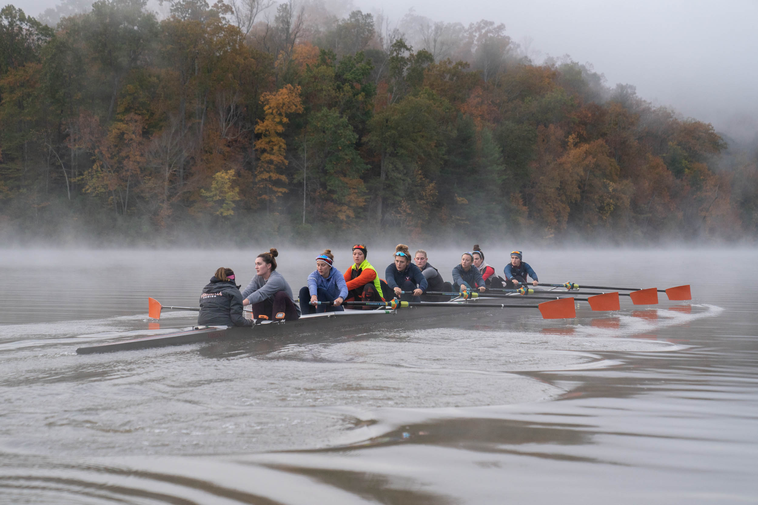 UVA womens rowing team on a foggy river with the bordering trees turning various fall colors