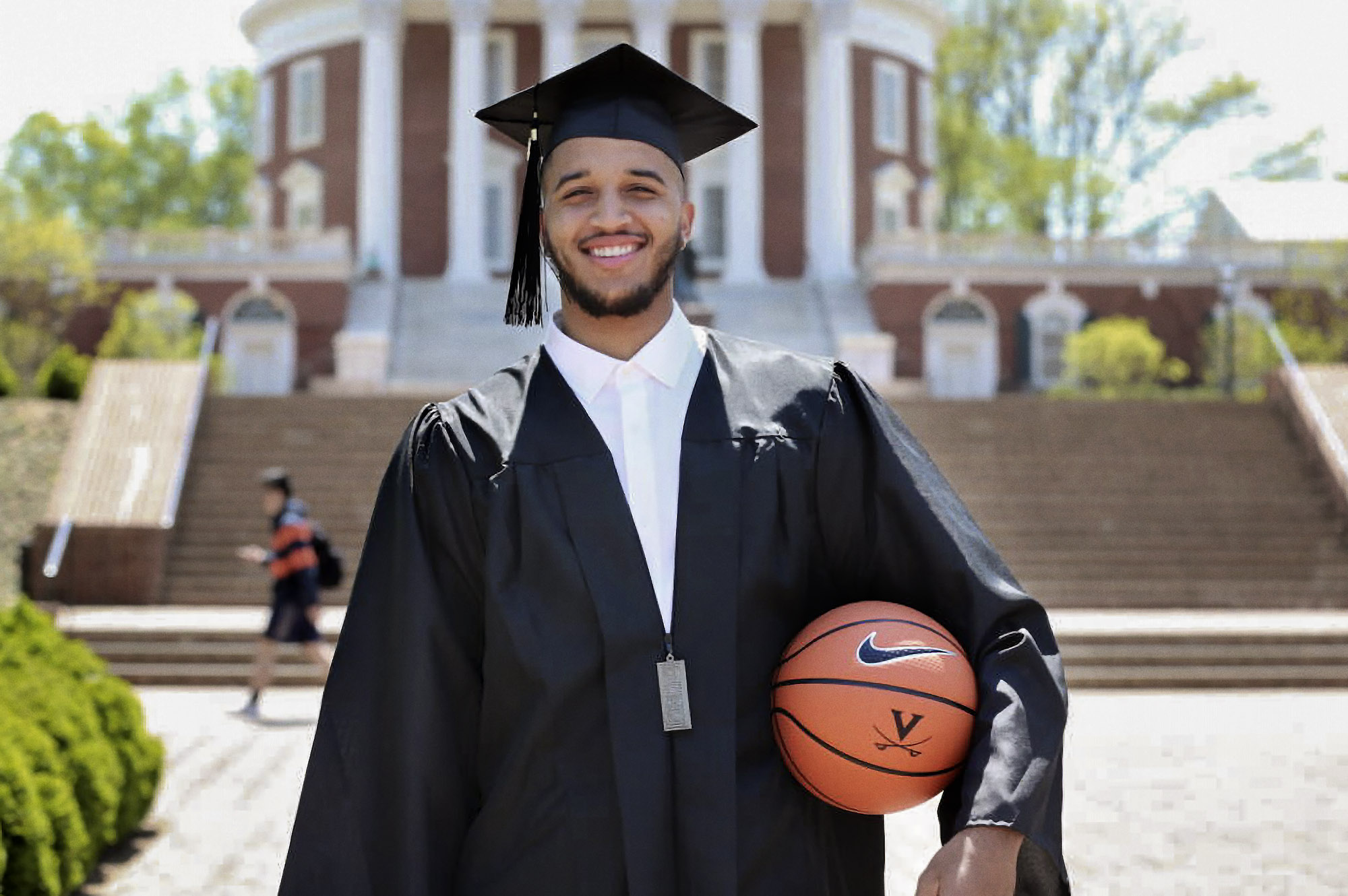 Isaiah Wilkins standing in graduation cap and gown holding a basketball in his left arm in front of the Rotunda for a picture