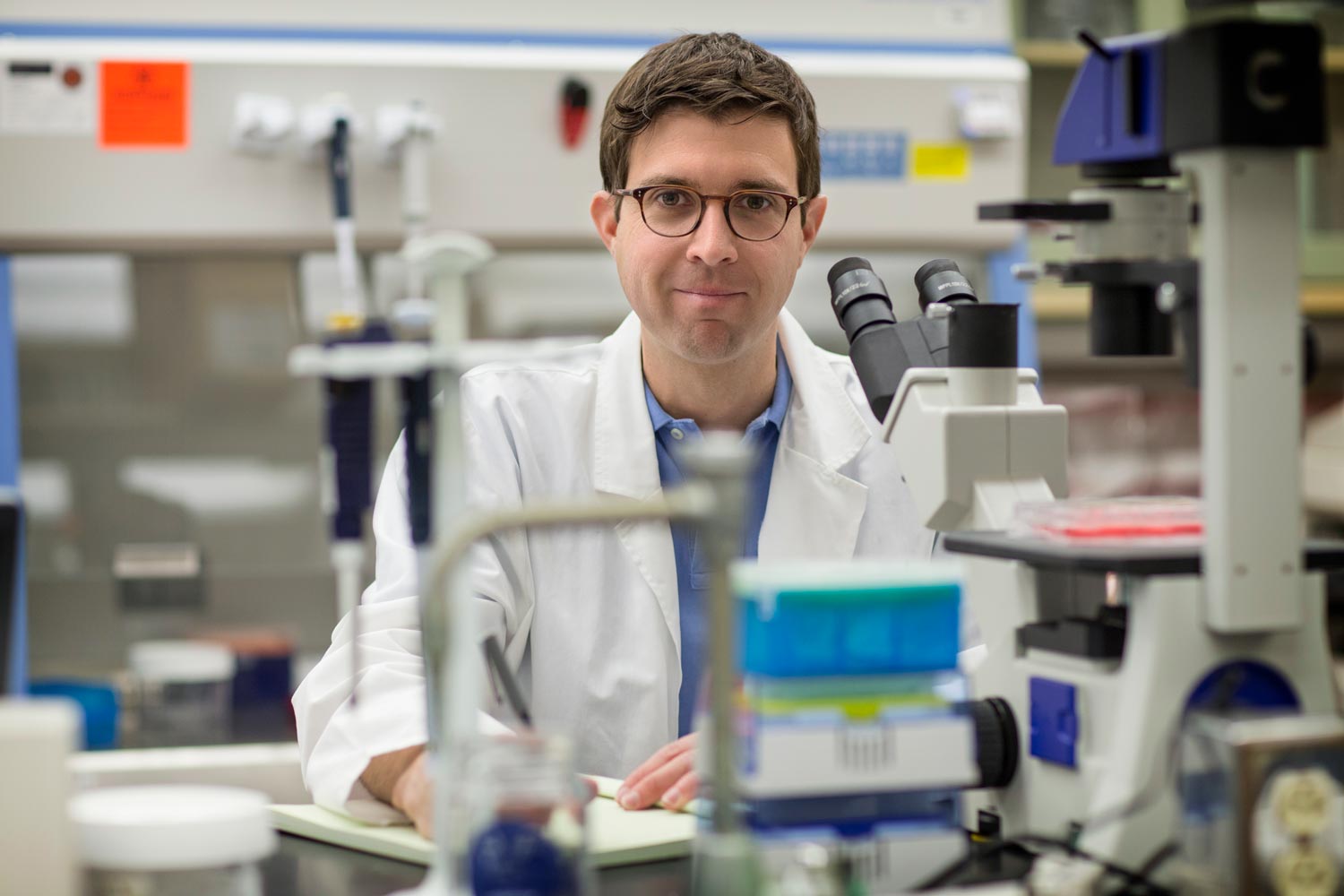 John Lukens sits at a microscope with pen and paper looking at the camera