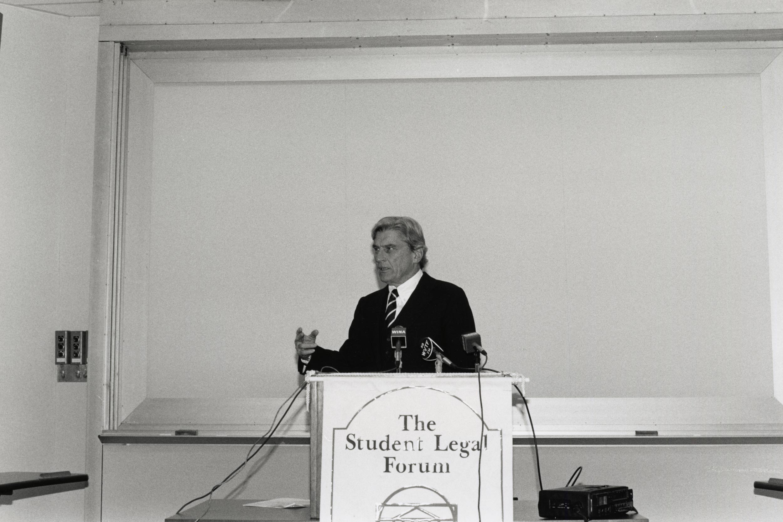 Black and white photo of John Warner speaking from a podium that reads The Student Legal Forum