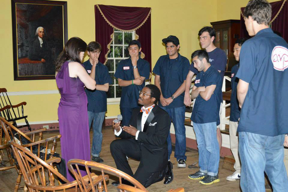 Man gets on one knee and proposes to a woman while his friends are gathered around waiting for her answer