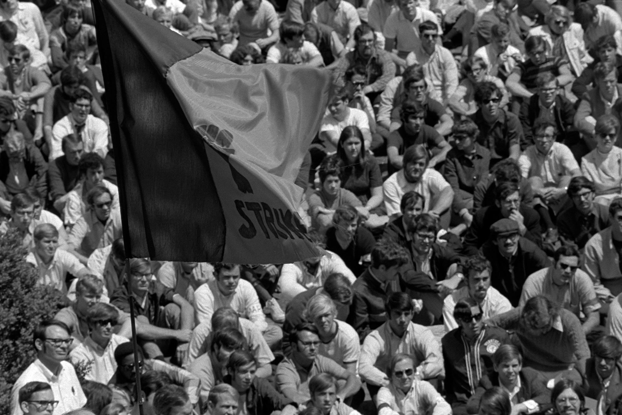 Black and White image of men sitting on the ground waving a two colored flag, but unable to know the colors or see the words on the flag
