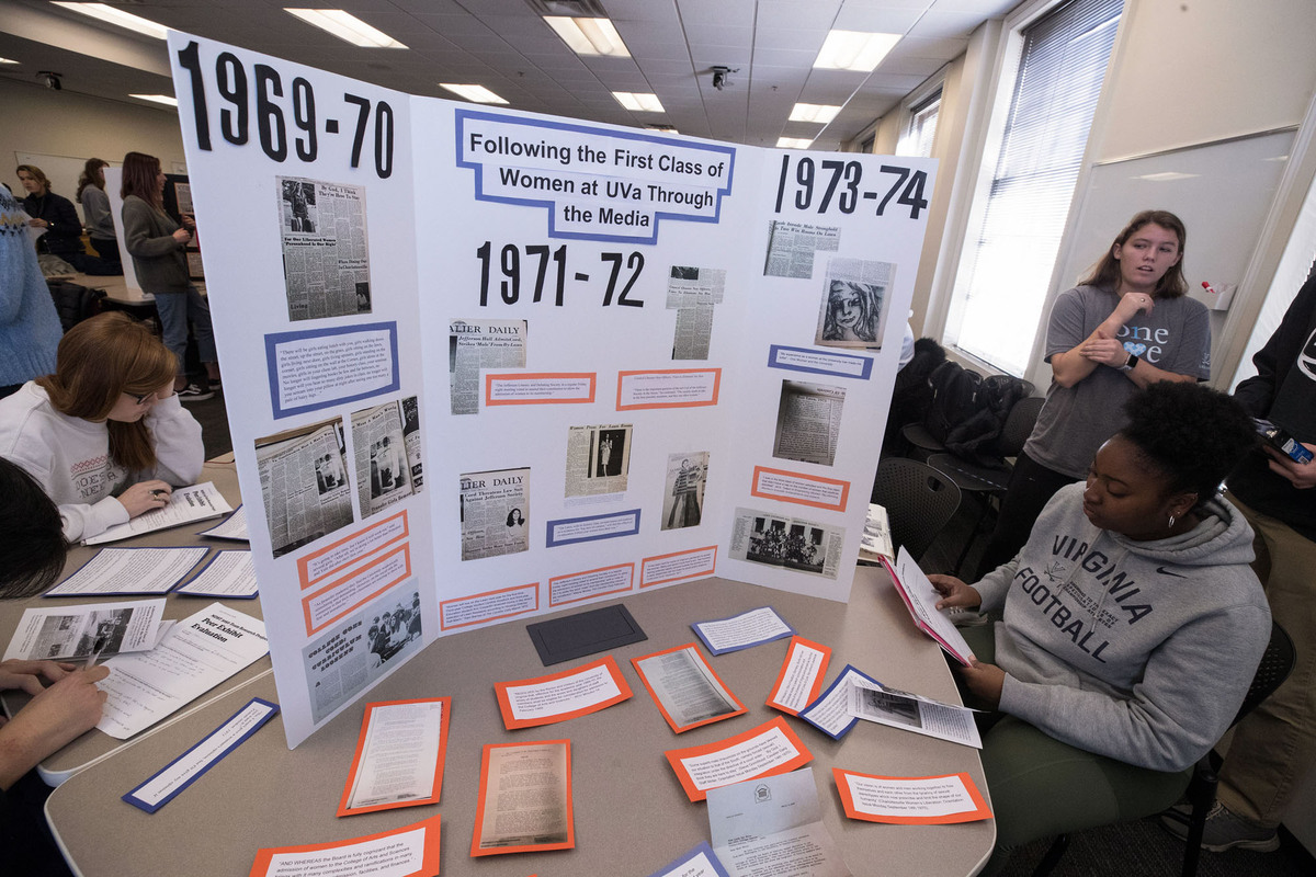 Woman sitting at a table with a tri-fold that is titled Following the First Class of Women at UVA through the Media