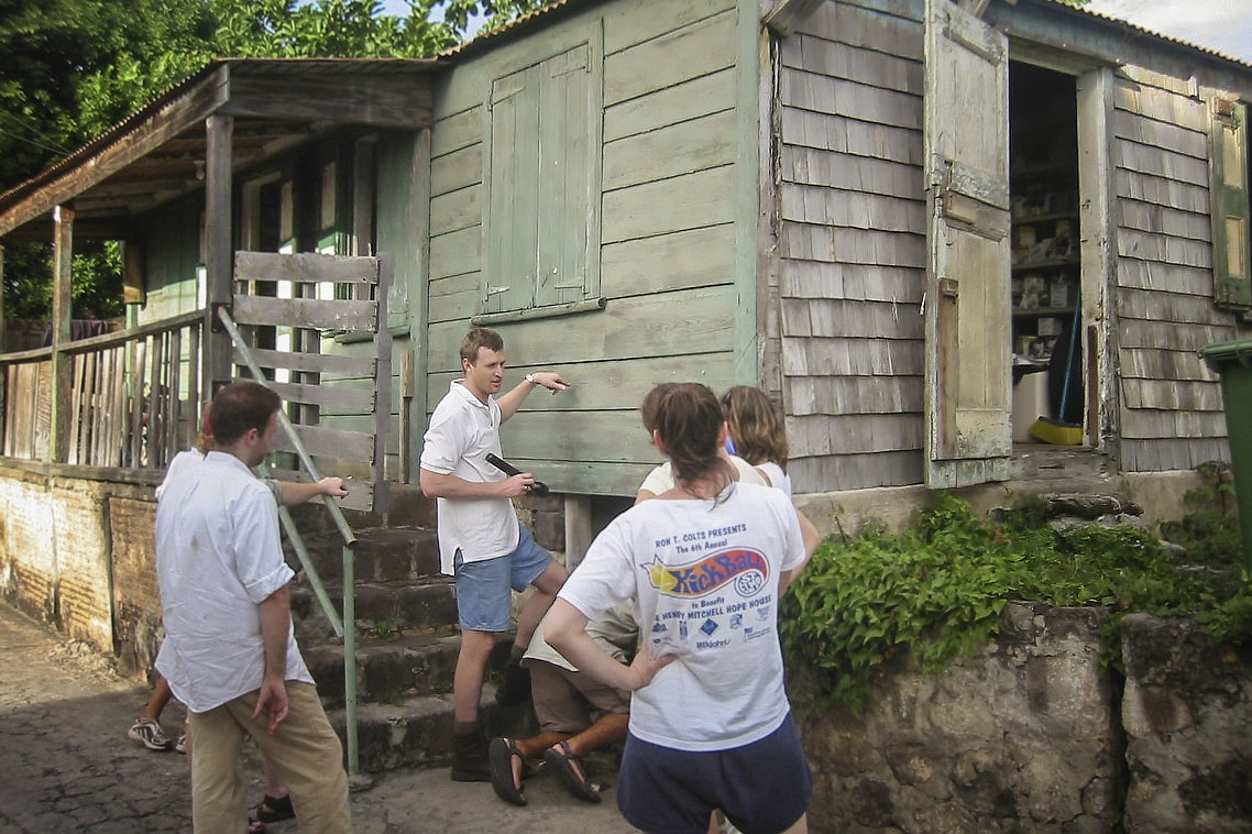 Louis Nelson holds a flashlight while he points to a worn down wooden building and talking to a group of students