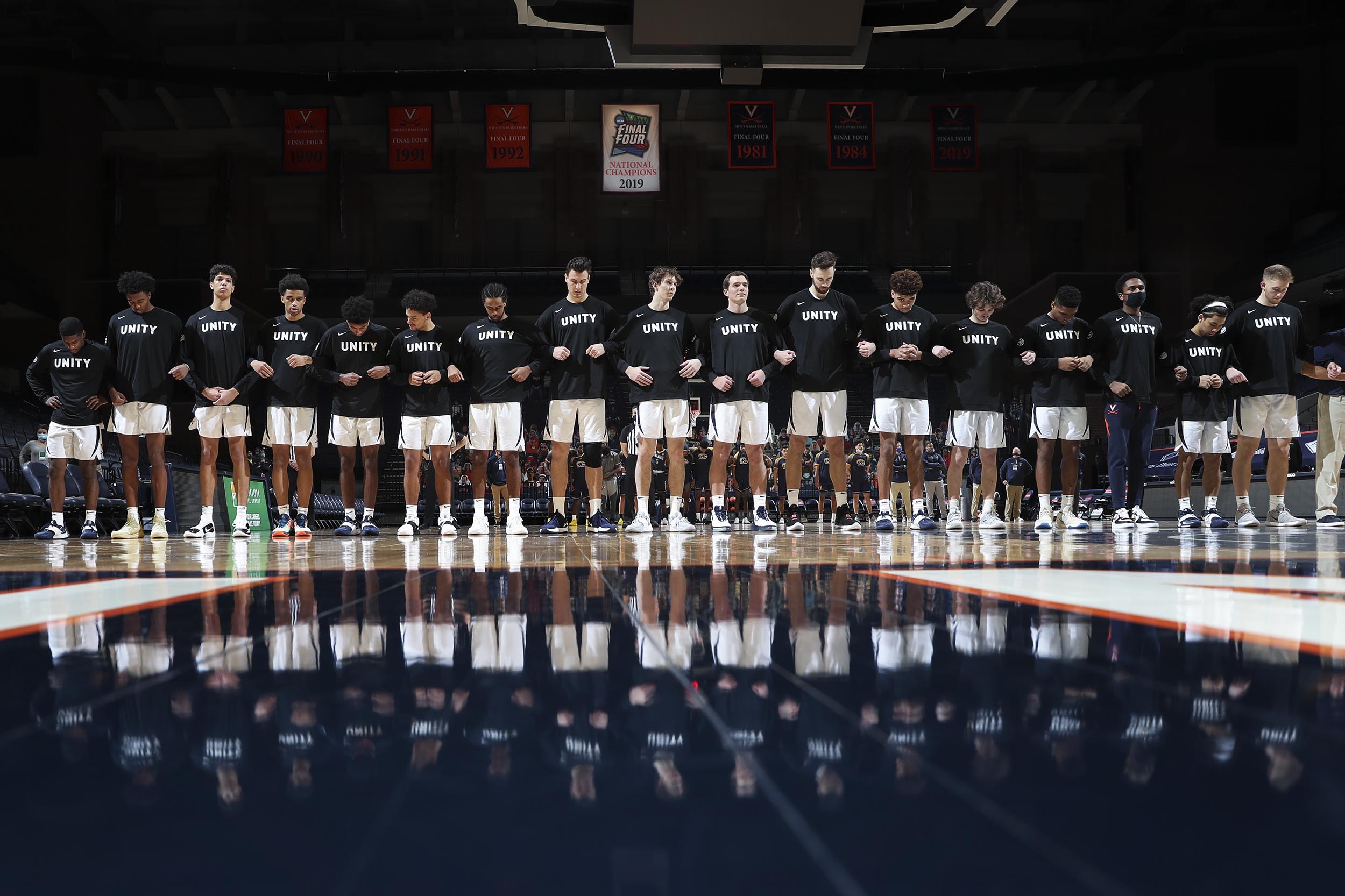 UVA mens basketball team interlock arms and look at the floor during the national anthem.  The whole team is wearing shirts that say unity.