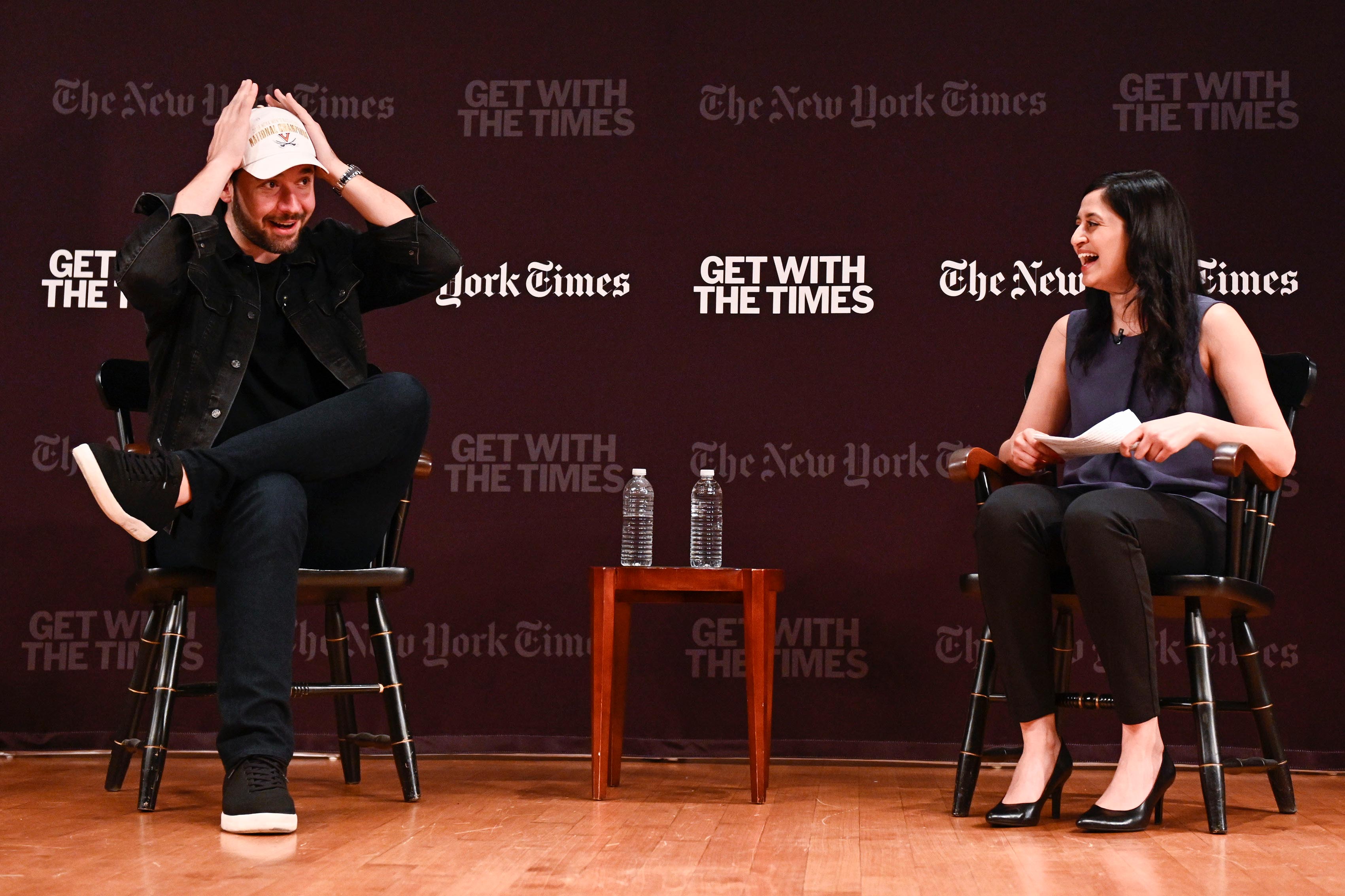 Ohanian, left, puts on a UVA national champions on stage while at a speaking engagement an woman sitting in a chair next to him is laughing