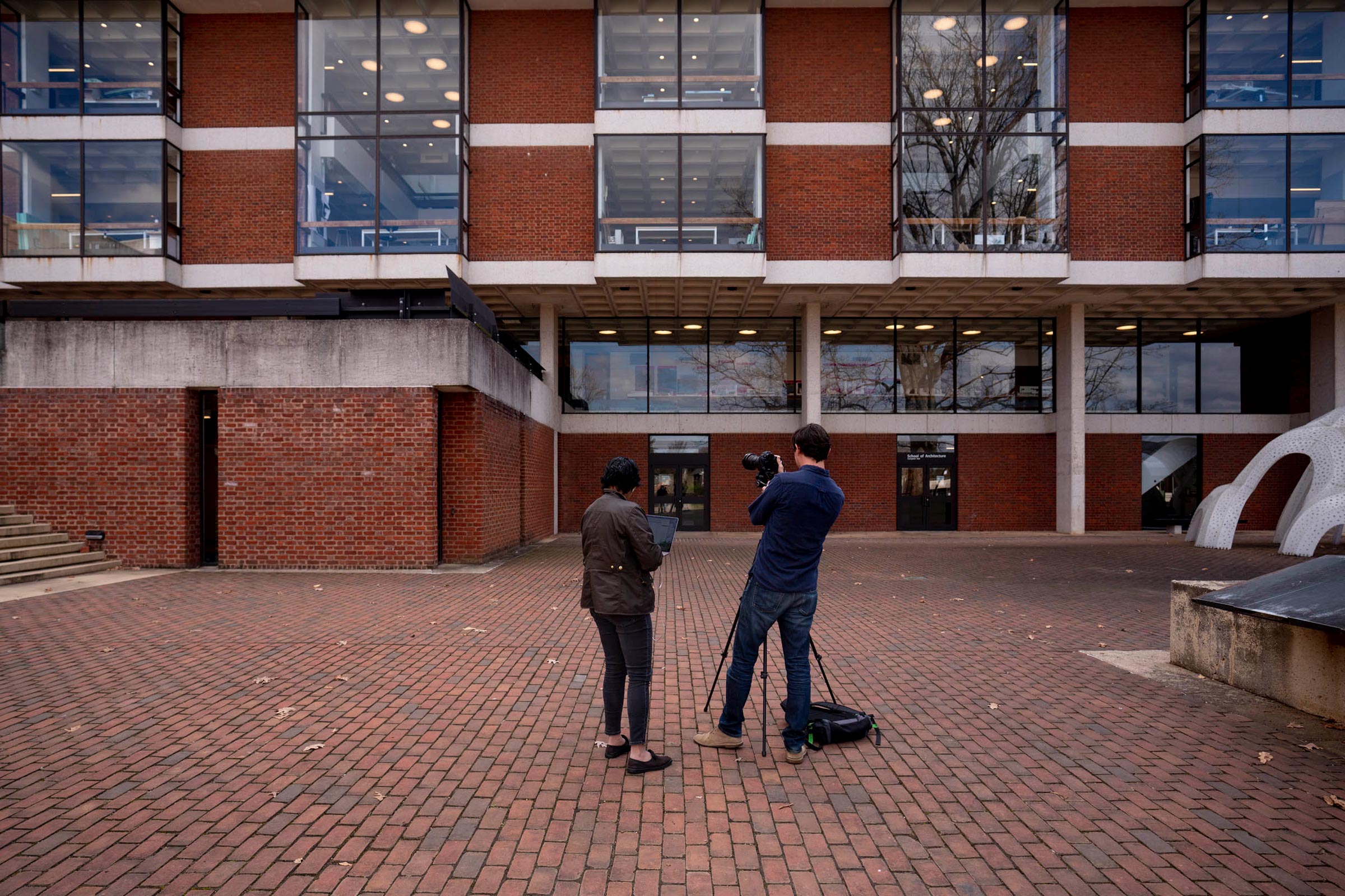 Two people standing outside of a big brick building.  One has a camera on a tripod taking pictures and one is holding a laptop