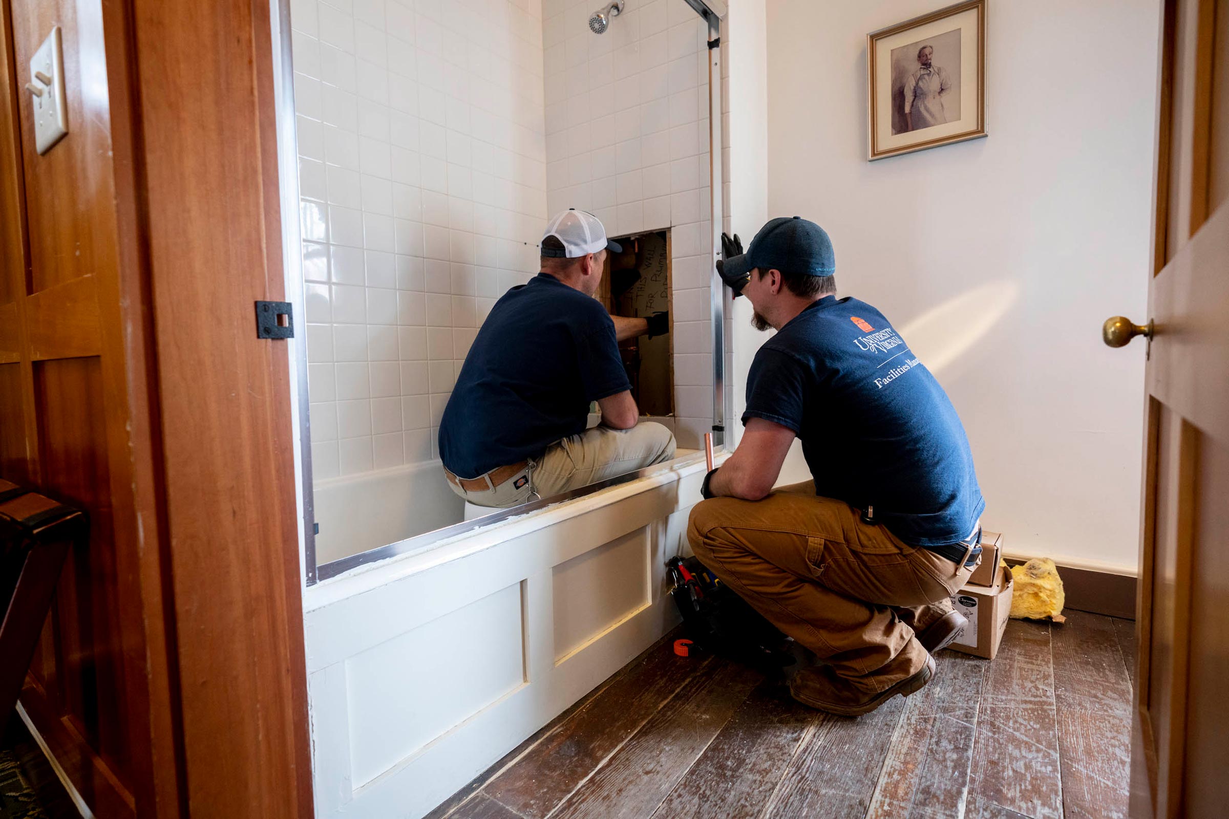 Two facilities management workers working on a bath tub.  One is sitting on a white 5 gallon bucket and looking into a whole in the shower wall and the second is bent down watching