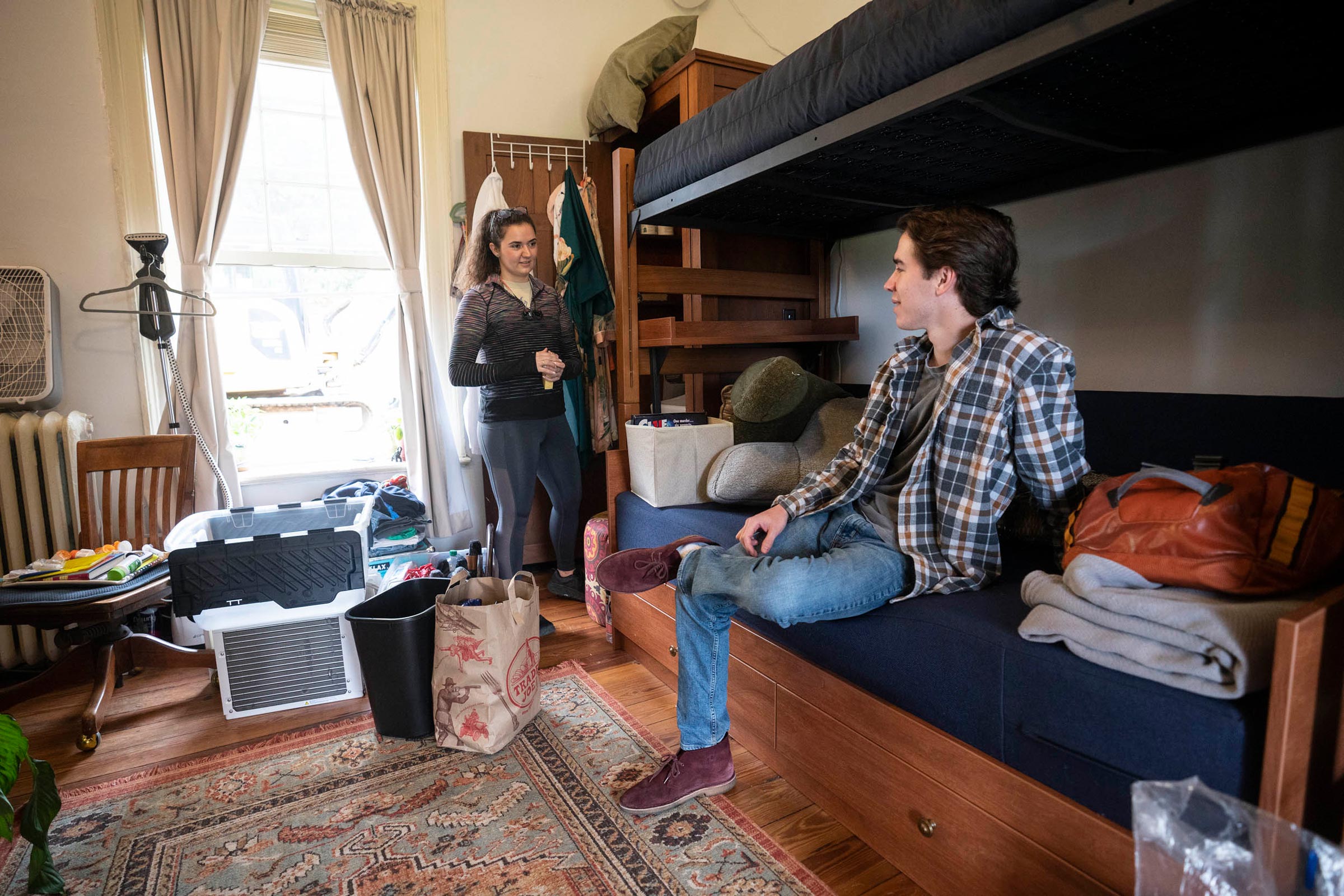 Two students in a dorm room talking.  Left person is standing at the closet right: person is sitting on couch