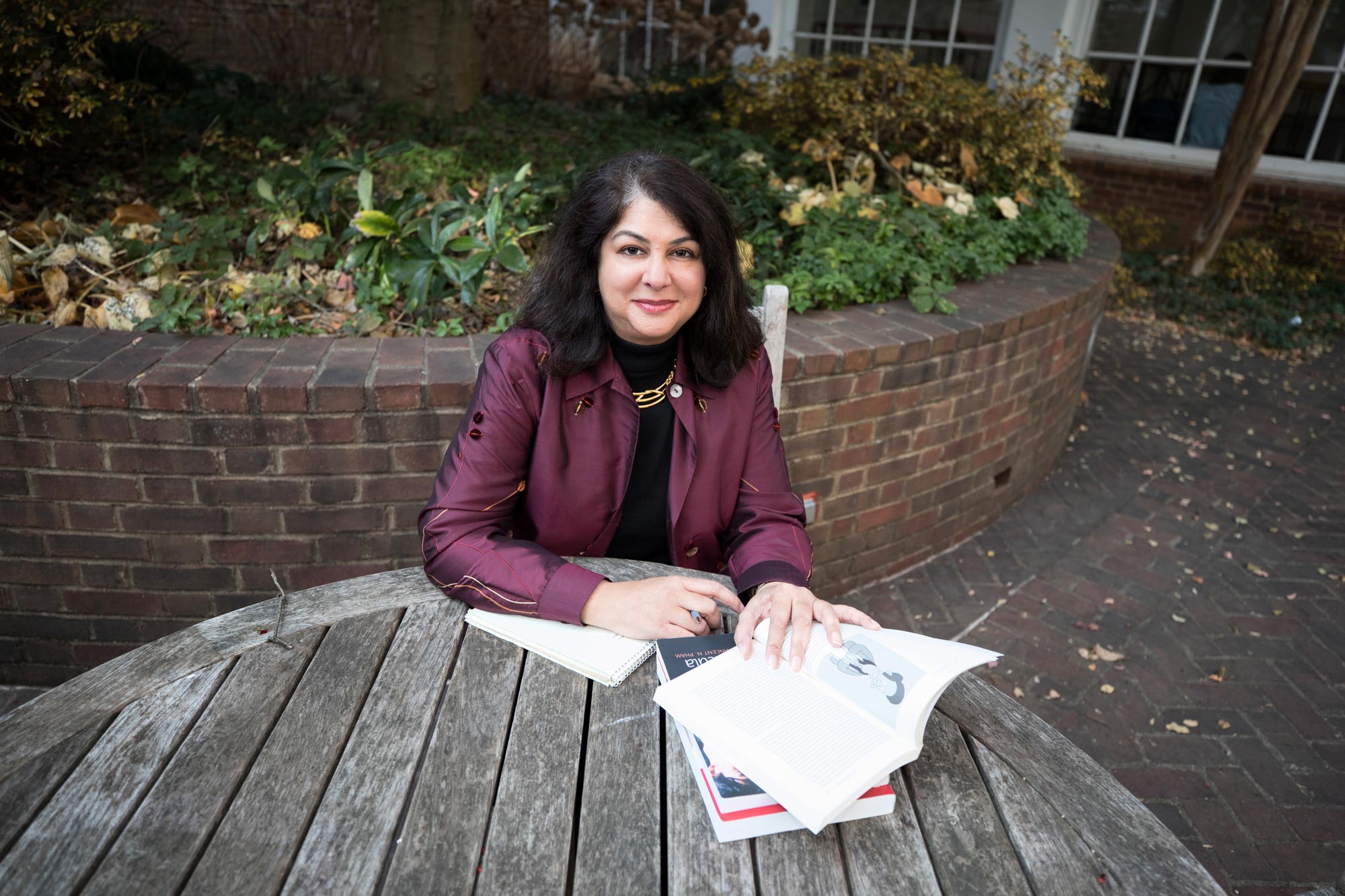 Shilpa Davé sitting at a circle wooden table looking at the camera with 4 books on the table with her left hand holding one open