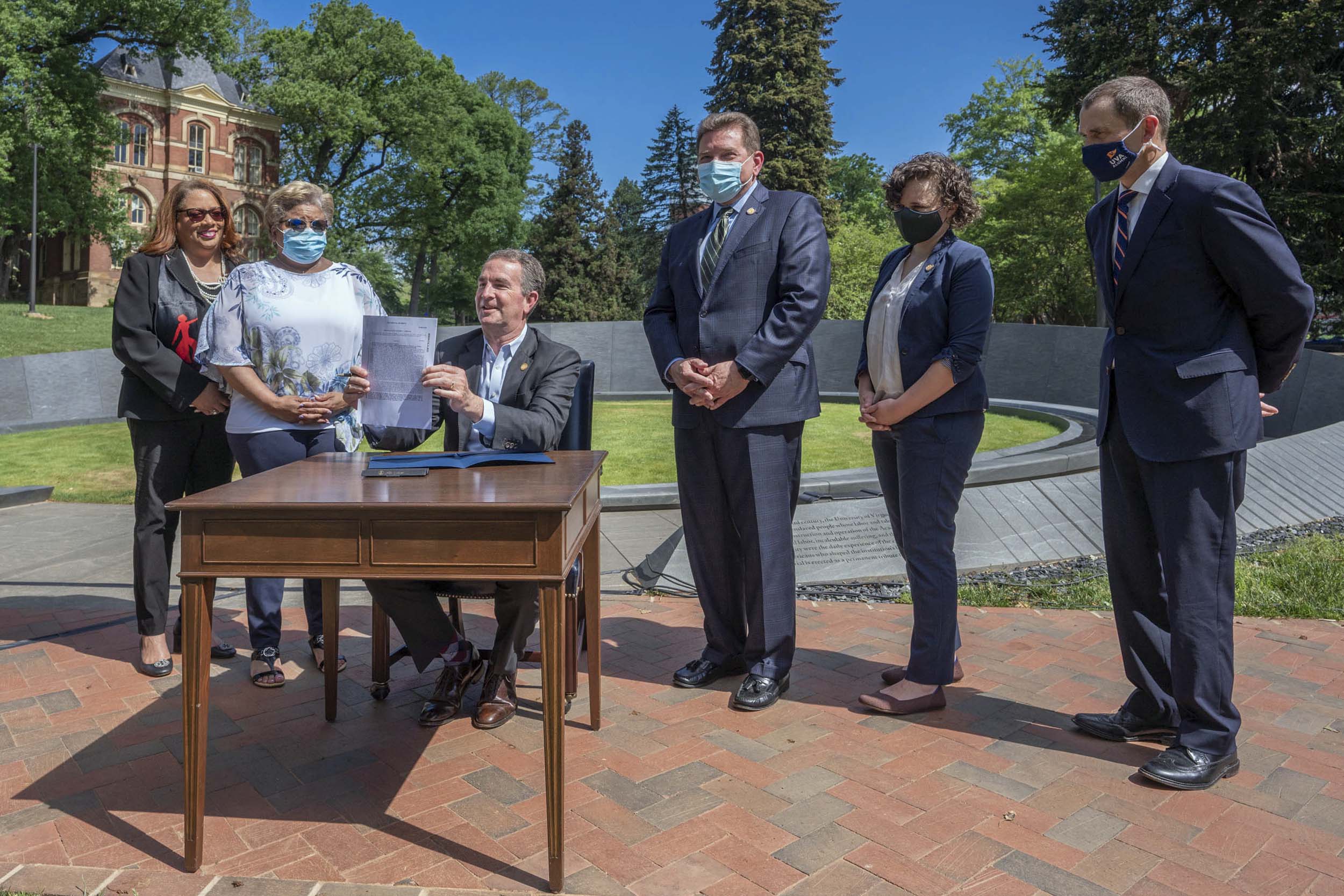 Ralph Northam sitting at a table holding up a piece of legislation at the Memorial for Enslaved Laborers while being surrounding by UVA members.