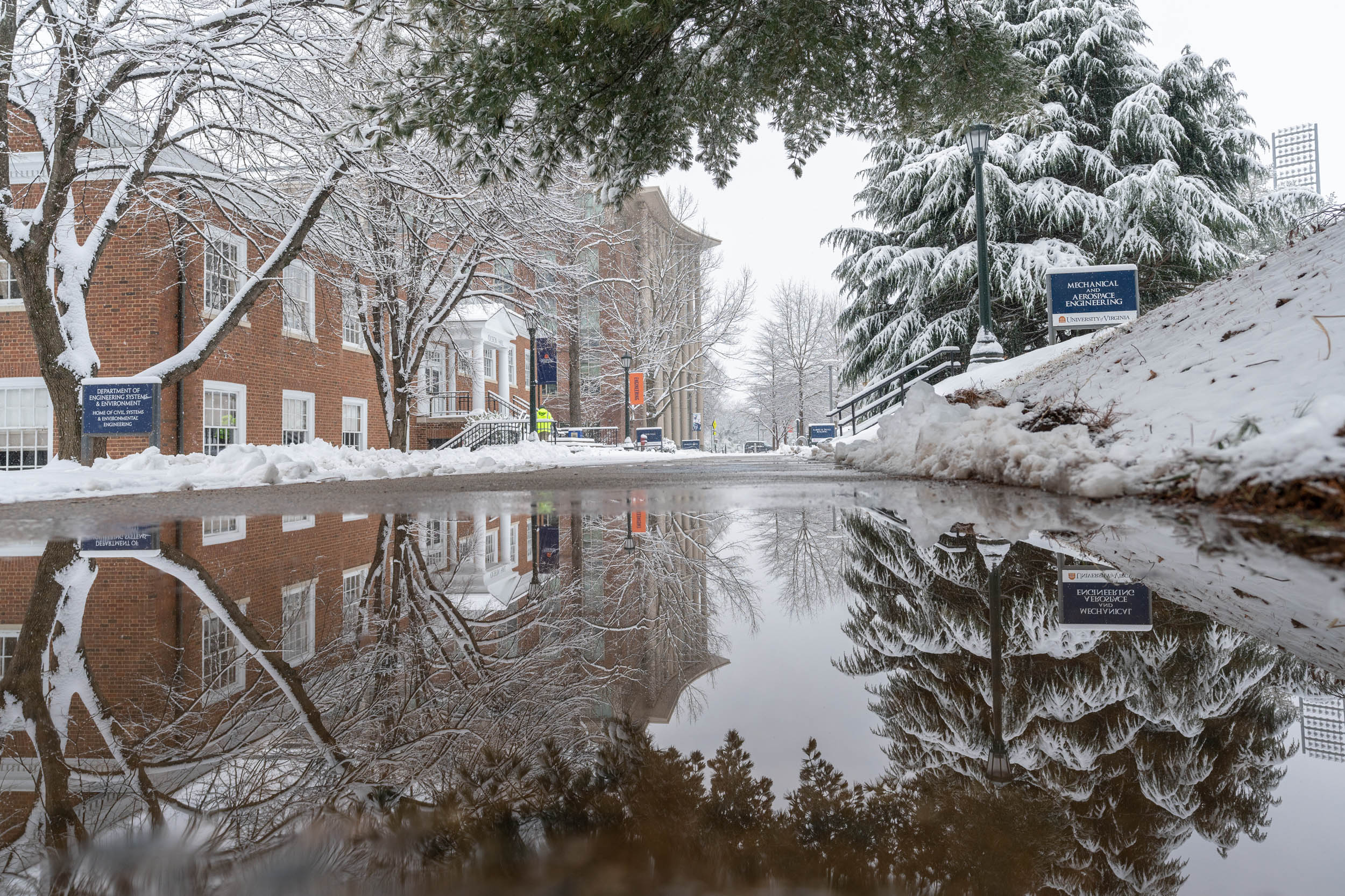 Snow covered trees and building reflecting off of a puddle on road