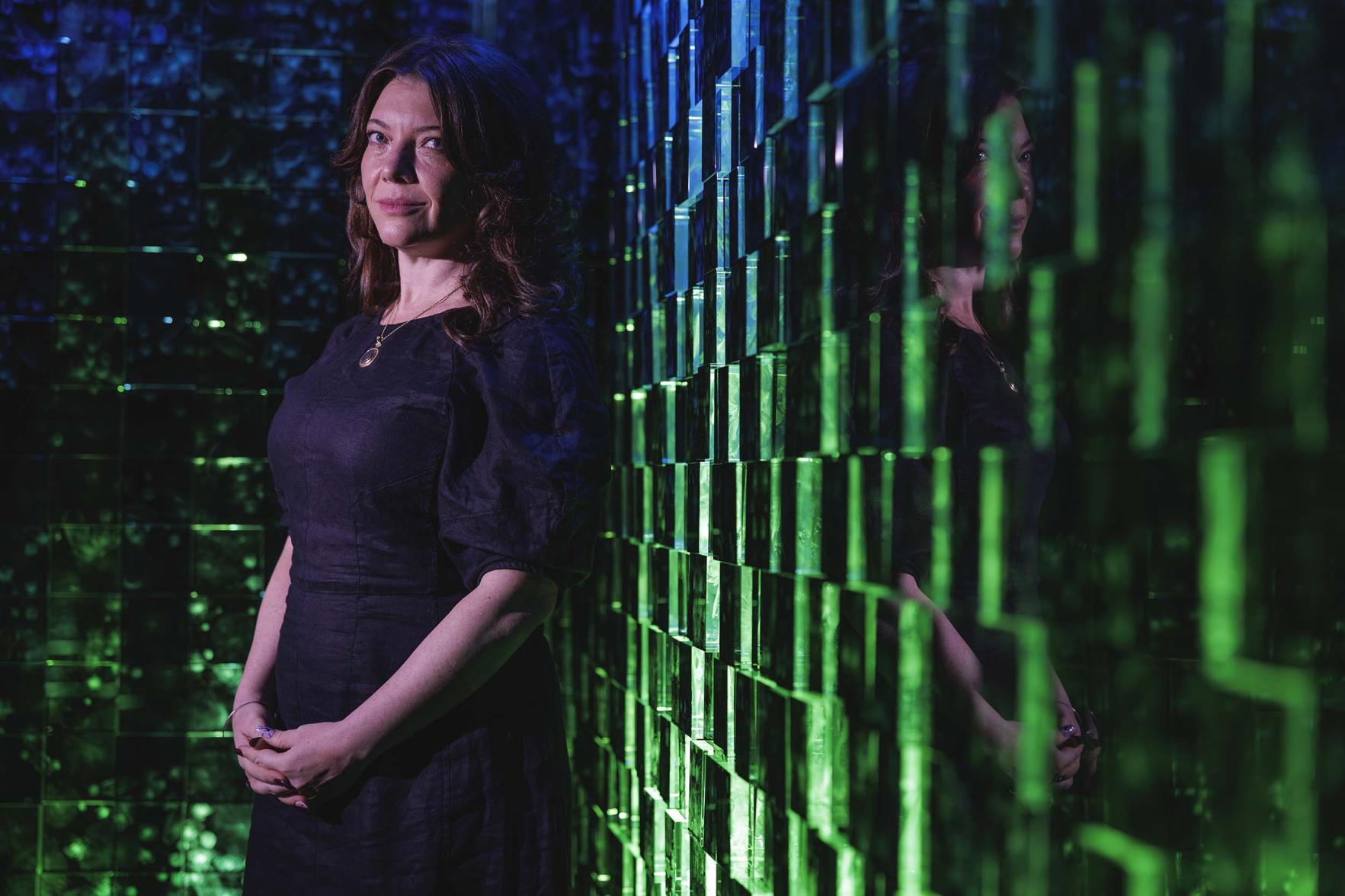 Lana Swartz standing against a glass sculpture wall in a dark, mysterious space.