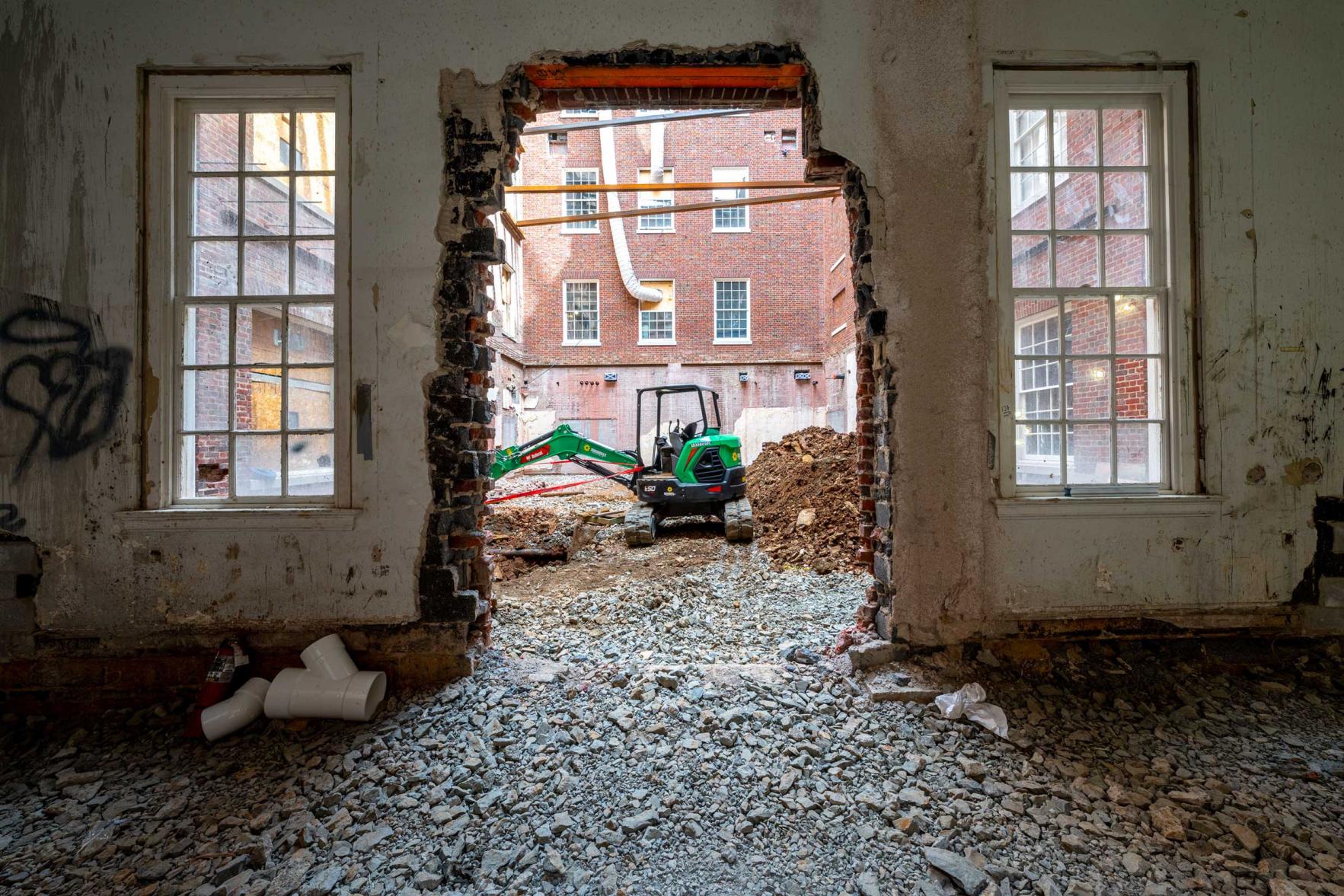 Through a demolished opening in a brick and plaster wall, a mini-excavator is visible in a courtyard