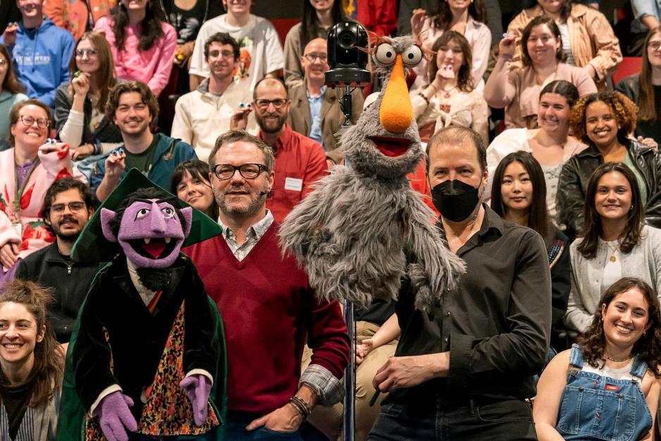 Two puppeteers hold Sesame Street characters, Count von Count and a gray fuzzy bird, in front of a cheering audience.