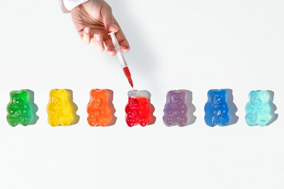 A row of different colored gummy bears with a hand holding a syringe filling one with red dye