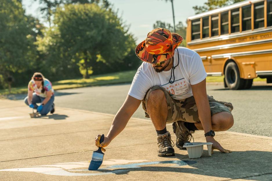 A volunteer squats to paint a white design on pavement near a school bus