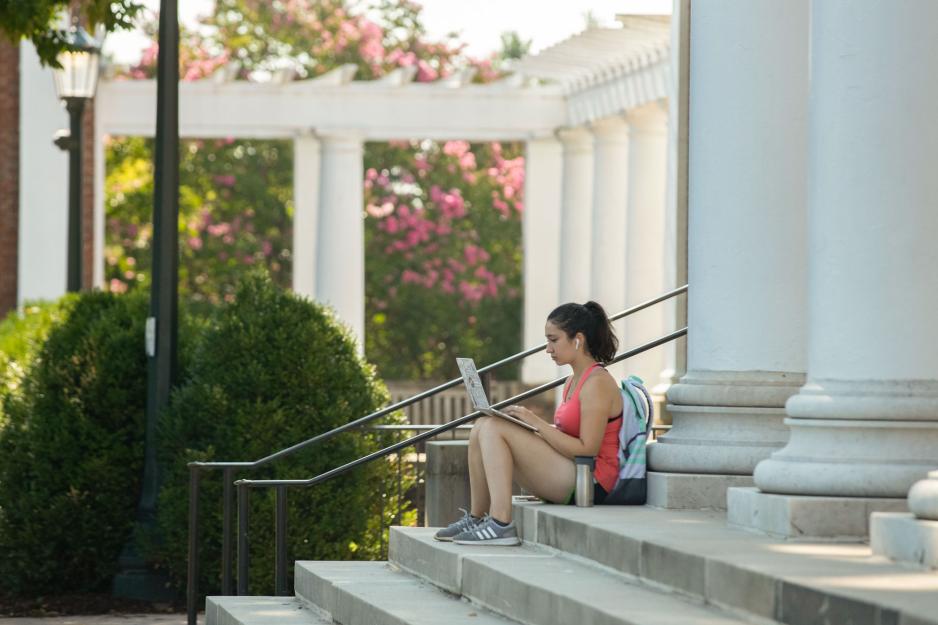 A UVA student wearing a backpack works on a laptop while seated on the steps of the Rotunda