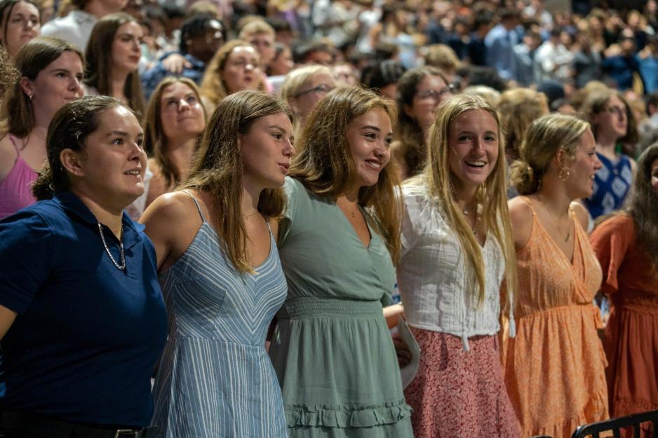 Five women with their arms around each other's backs singing in the crowded auditorium