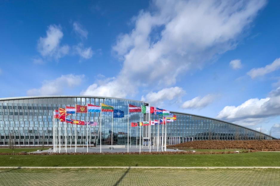 Flags of NATO member countries forms a circle outside the glass NATO headquarters building