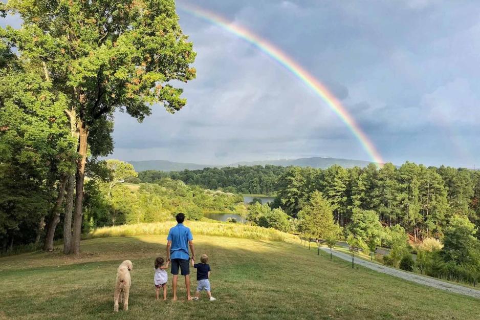 Father, two kids, and the family dog stand on a hill looking up at a rainbow