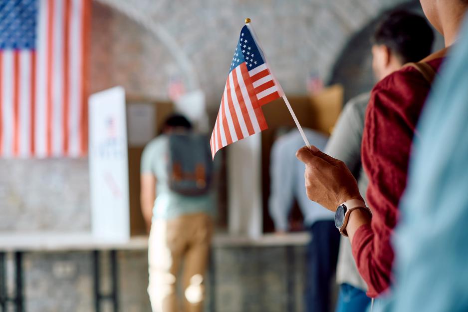 A blurred image of two people in voter booths with a line of three people waiting their turn, one holding an American flag.