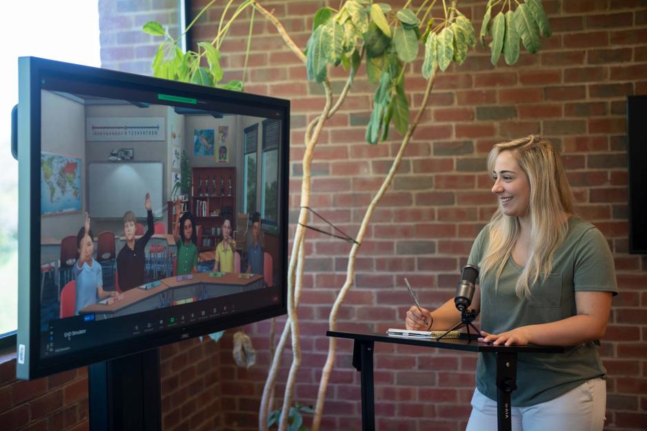 A woman stands at a podium, next to a large screen displaying a Zoom meeting with computer generated children raising their hands