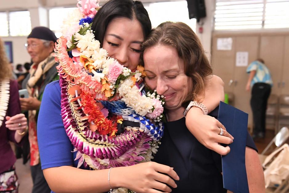 Esther Kwon coverd in ceremonial leis hugging a colleague in her elementary school